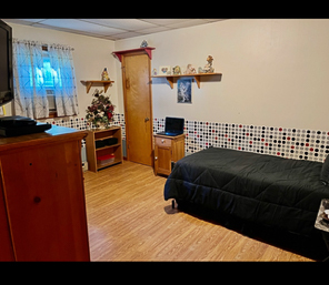 A bedroom with wood flooring, a twin bed with a black comforter, a wooden dresser, a small desk with a laptop, and shelves displaying decorations above a patterned accent wall inside the home of Sponsored Residential Providers Beth and Otis Fowler in Rustburg, Virginia.