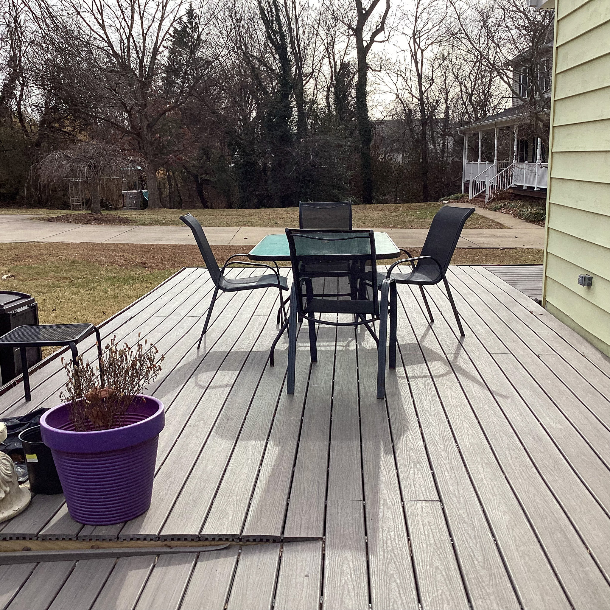 Outdoor wooden deck with a glass-top table, four black chairs, and a potted plant overlooking a grassy yard inside the home of Sponsored Residential Provider Eglee Escorihuela.