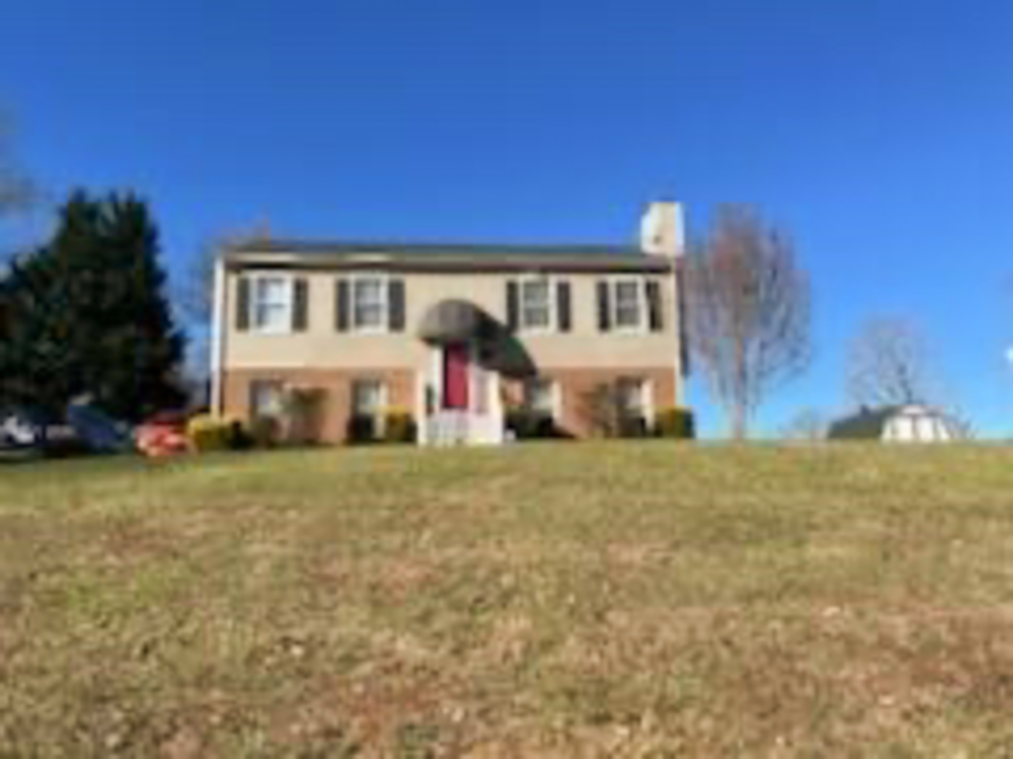 A two‑story house with tan siding, dark shutters, and a red front door sits on a sloped lawn under a clear blue sky belonging to Sponsored Residential Provider Devonne Dixon in Lynchburg, Virginia.