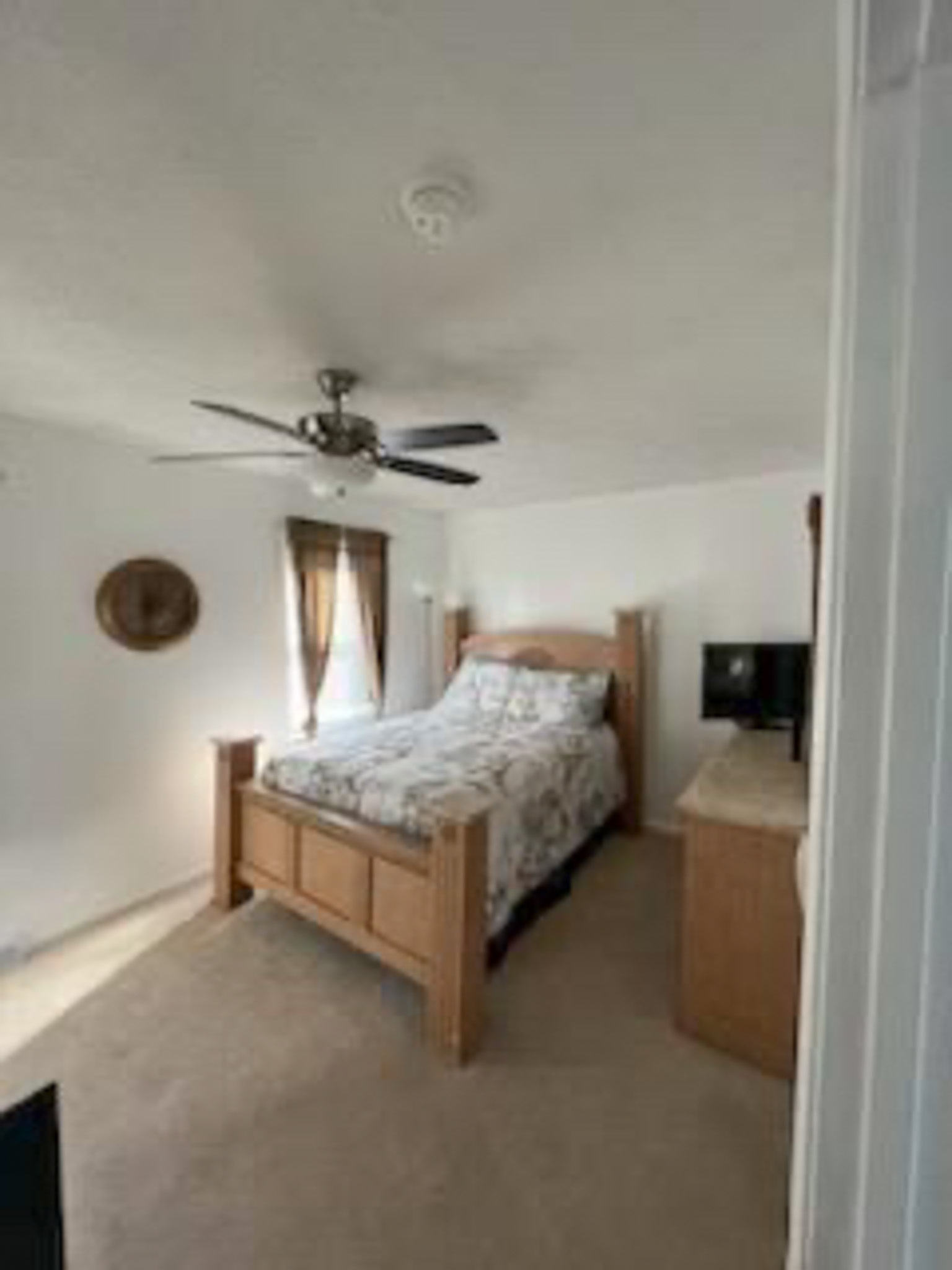 A bedroom with a light wood bed frame, patterned bedding, a ceiling fan, carpeted floors, and a matching dresser beside the bed inside the home of Sponsored Residential Provider Devonne Dixon in Lynchburg, Virginia.