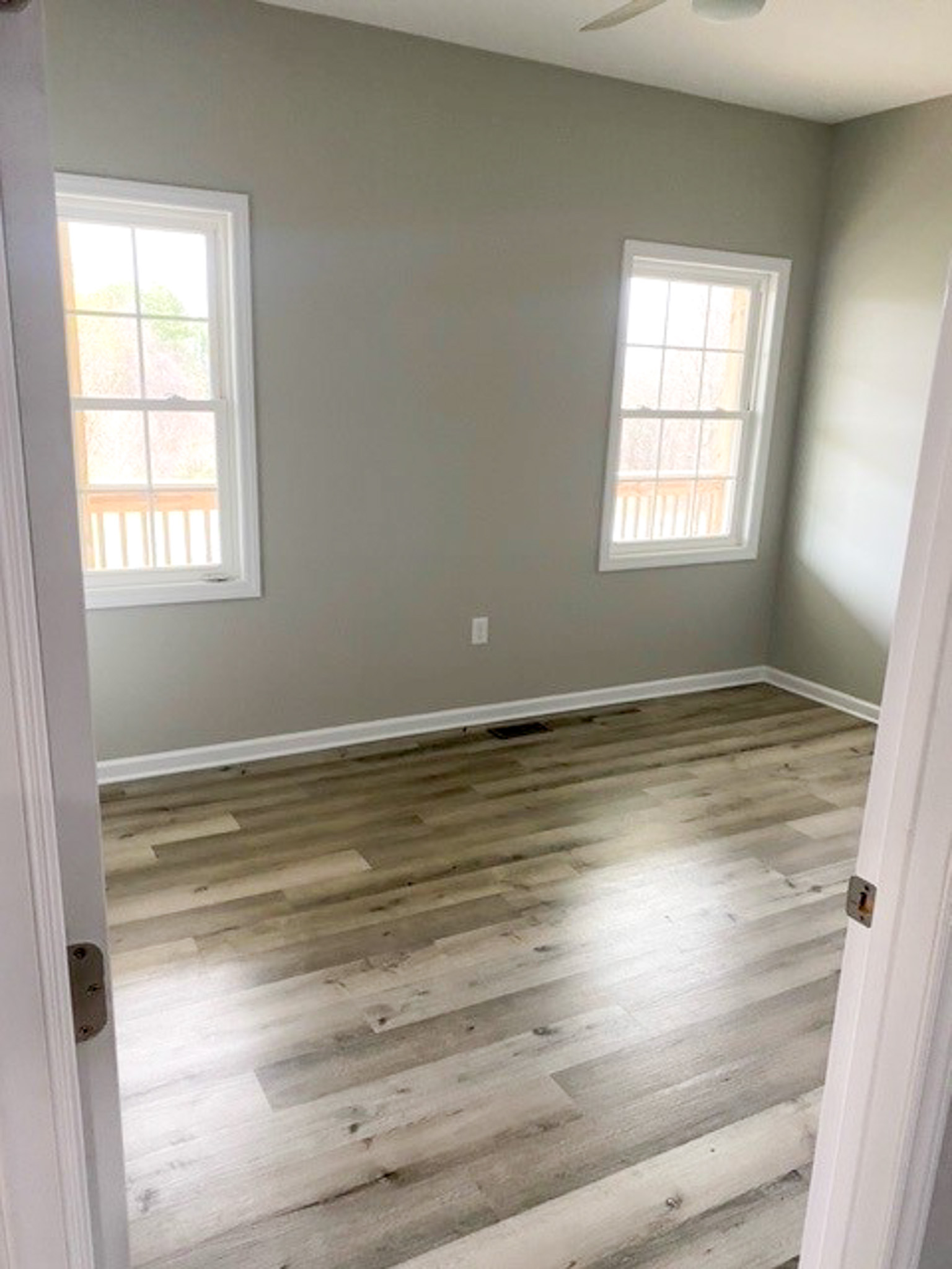 A bright, unfurnished bedroom inside Jake and Angie Beamer’s provider group home in Hillsville, Virginia, with two windows, neutral gray walls, and wood-look flooring.