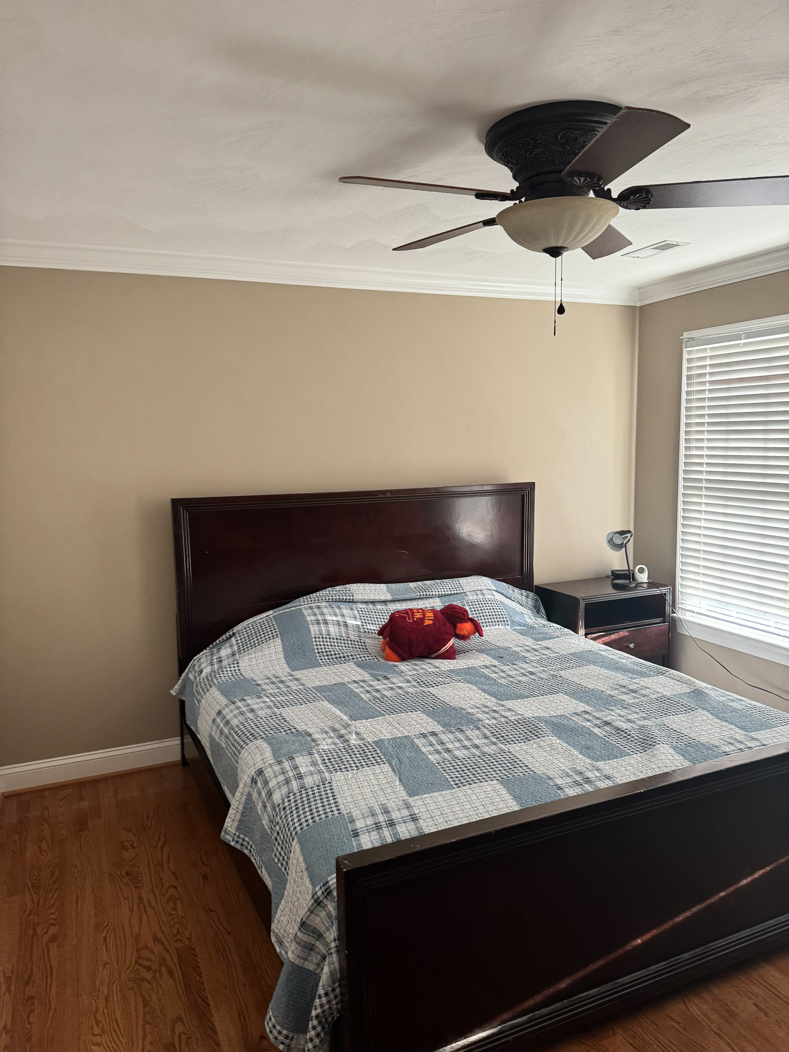 A bedroom with a dark wooden bed frame, a blue‑and‑white quilt, a ceiling fan, and light from a window with blinds inside the home of Sponsored Residential Provider Sharon Jennings in Salem, Virginia.