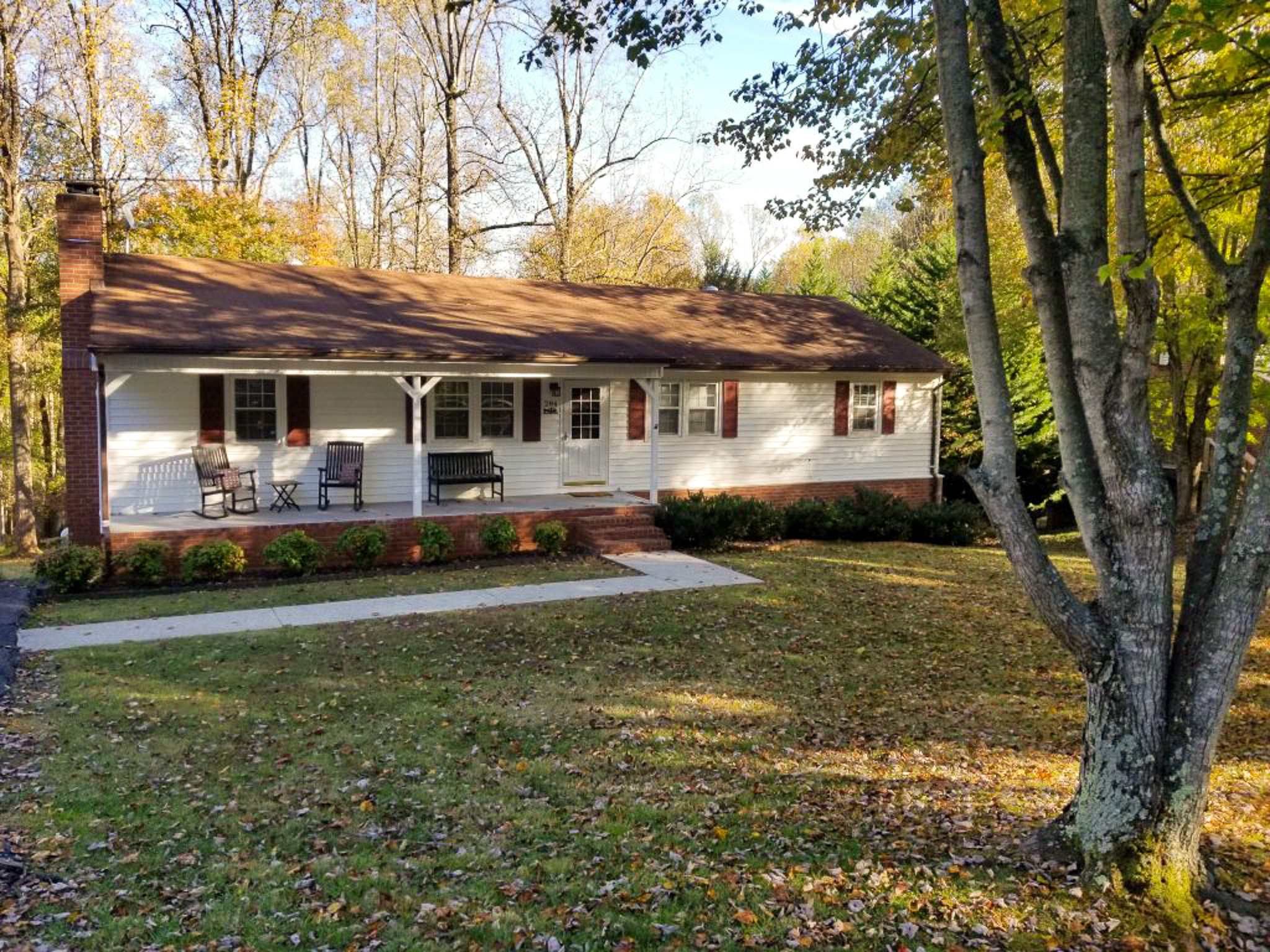 Single‑story white house with red shutters, a small front porch, and a large leaf‑covered yard bordered by tall trees belonging to Sponsored Residential Provider Kathi Evans in Lynchburg, Virginia.