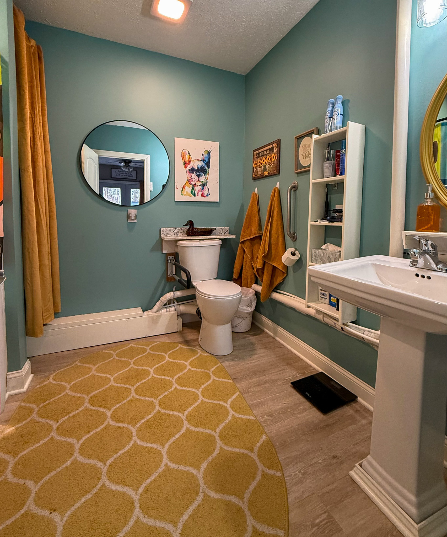 A bathroom with teal walls, a pedestal sink, a toilet, mustard-yellow towels, a round mirror, wall art, open shelving, and a yellow patterned rug inside the home of Sponsored Residential Providers Kim and Rick Duncan in Roanoke, VA.