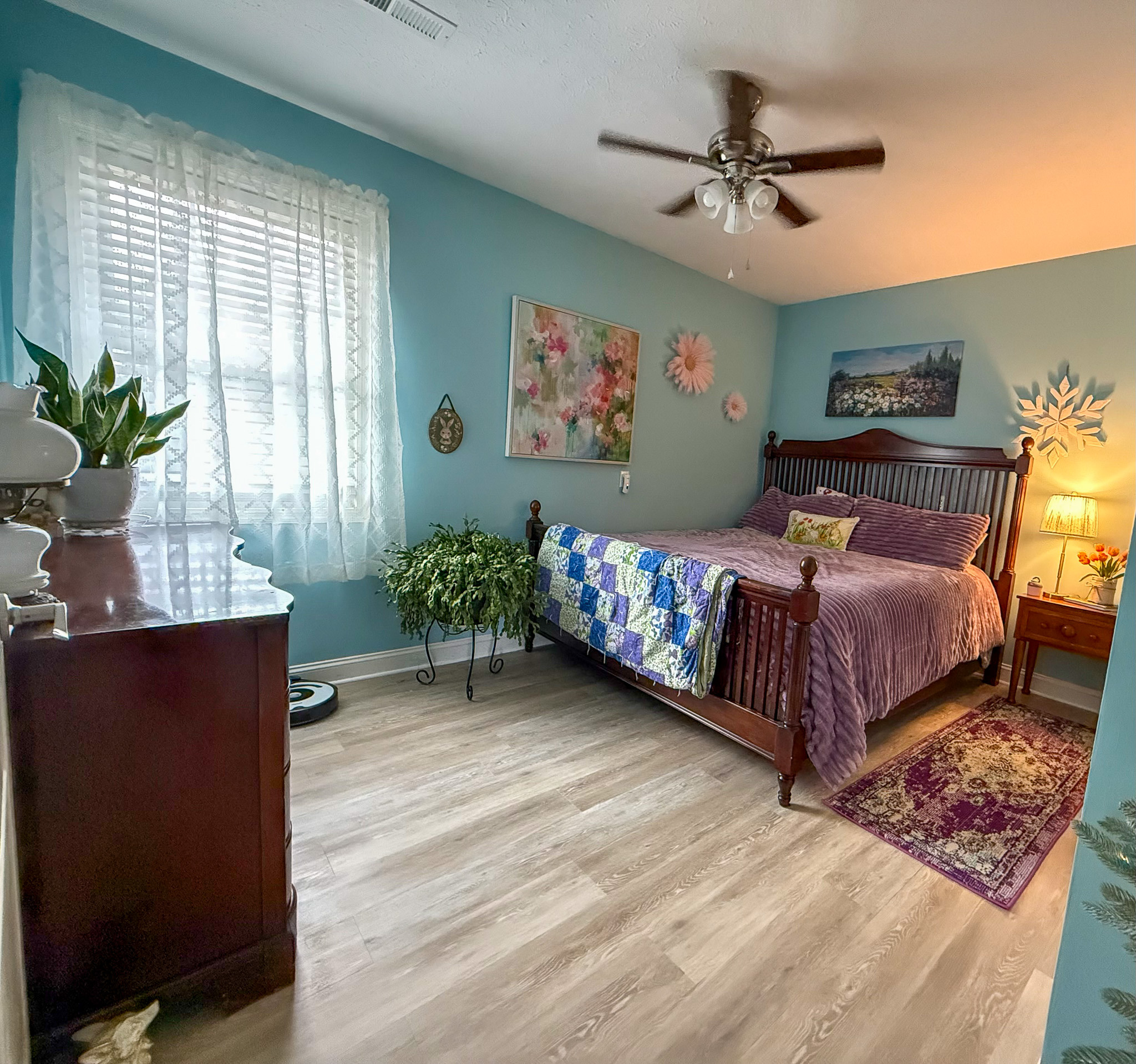 A cozy bedroom with light blue walls, a wooden bed with purple bedding, a quilt at the foot, plants, wall art, a wooden dresser, and a ceiling fan inside the home of Sponsored Residential Providers Kim and Rick Duncan in Roanoke, VA.