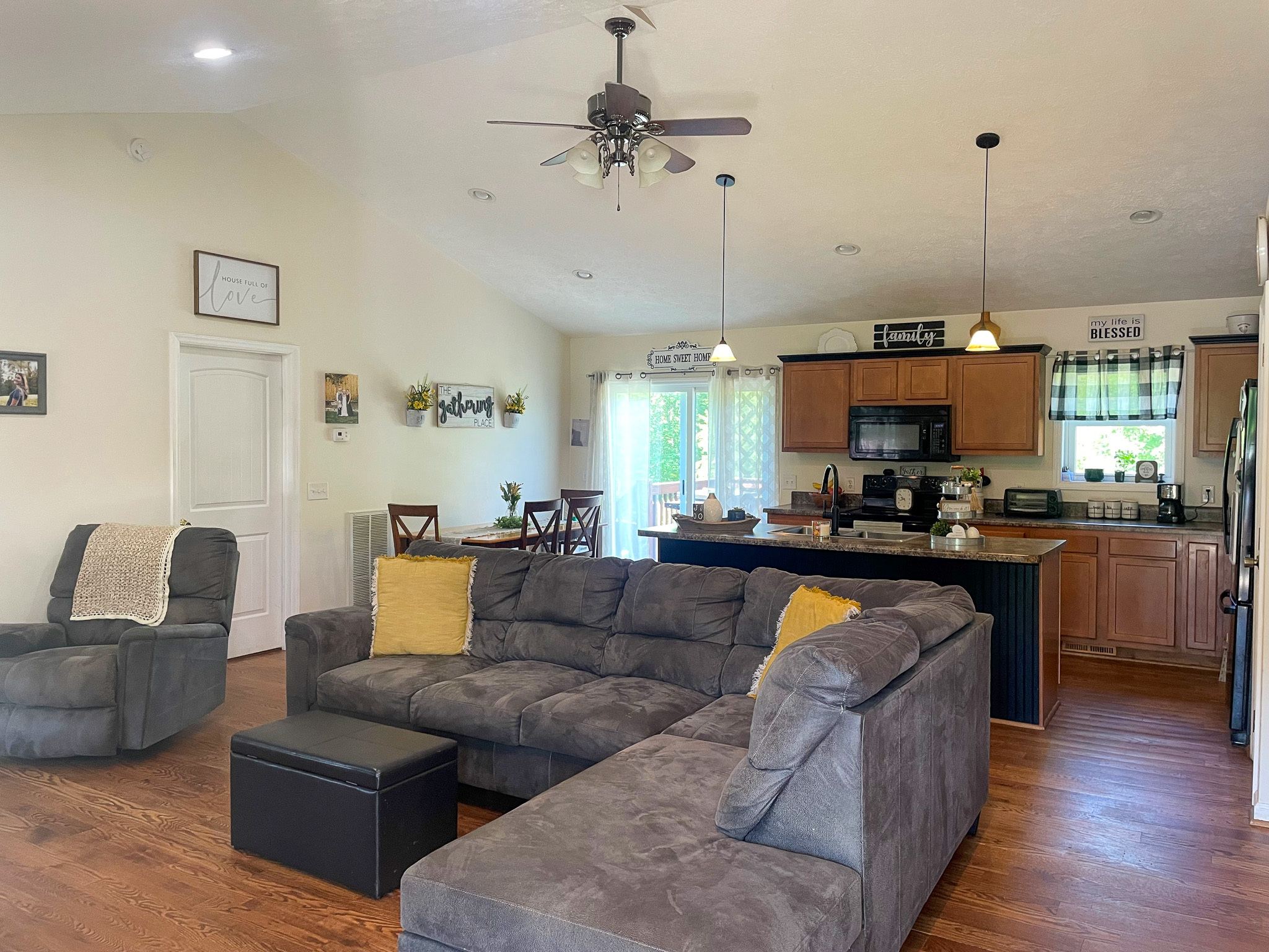 Open living area with a gray sectional sofa, recliner, ceiling fan, and a kitchen with wooden cabinets and a dining table in the background inside the home of Sponsored Residential Provider Chancie Haga in Woodlawn, Virginia.