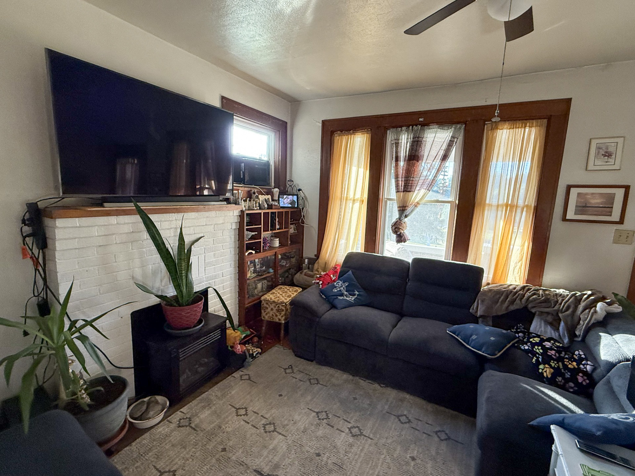 Living room with a white brick fireplace, large wall‑mounted TV, dark sofas, potted plants, and tall windows with sheer curtains inside the home of Sponsored Residential Provider Tammy Hunt in Floyd, Virginia.
