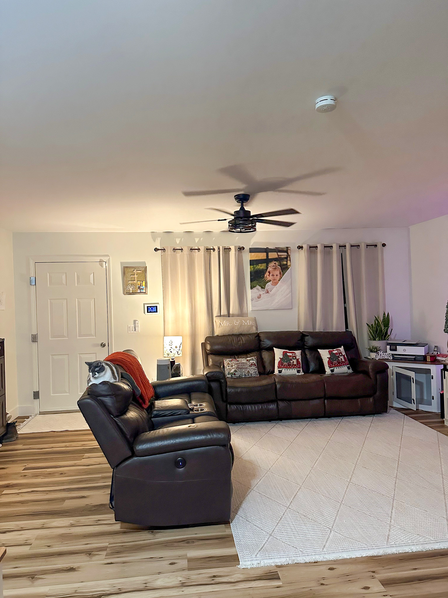 Living room with dark leather reclining sofas, a ceiling fan, light-colored curtains, hardwood flooring, a white area rug, and a small media stand with decor inside the home of  Sponsored Residential Provider Gabrielle Straway in Grottoes, Virginia.