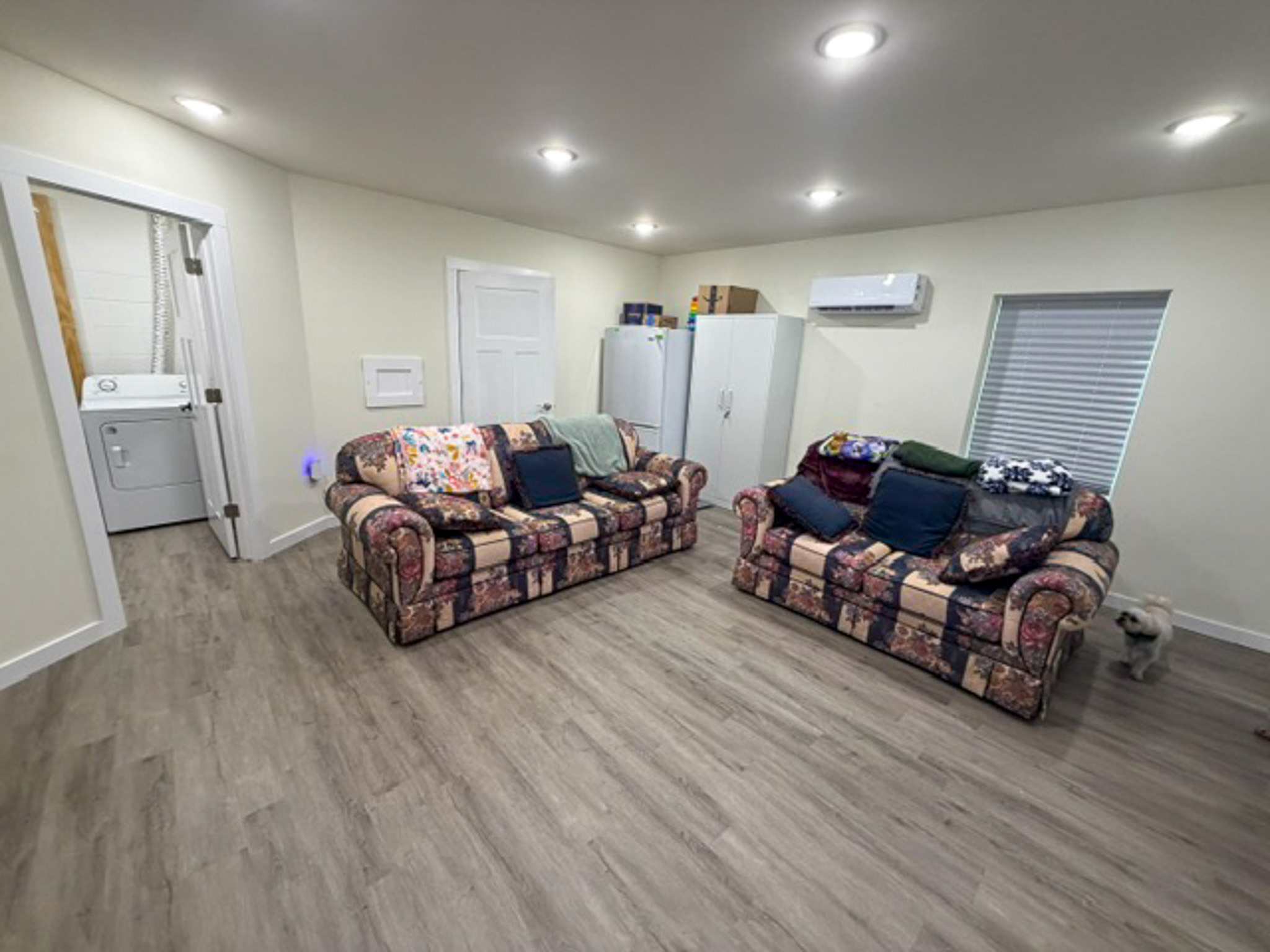 Spacious living room with gray wood-look flooring, recessed ceiling lights, and two patterned sofas facing each other. A laundry area is visible through an open doorway, with storage cabinets, a window with blinds, and a wall-mounted mini-split air conditioning unit along the back wall inside the home of Group Home Provider Luke Haynes in Christiansburg, VA.