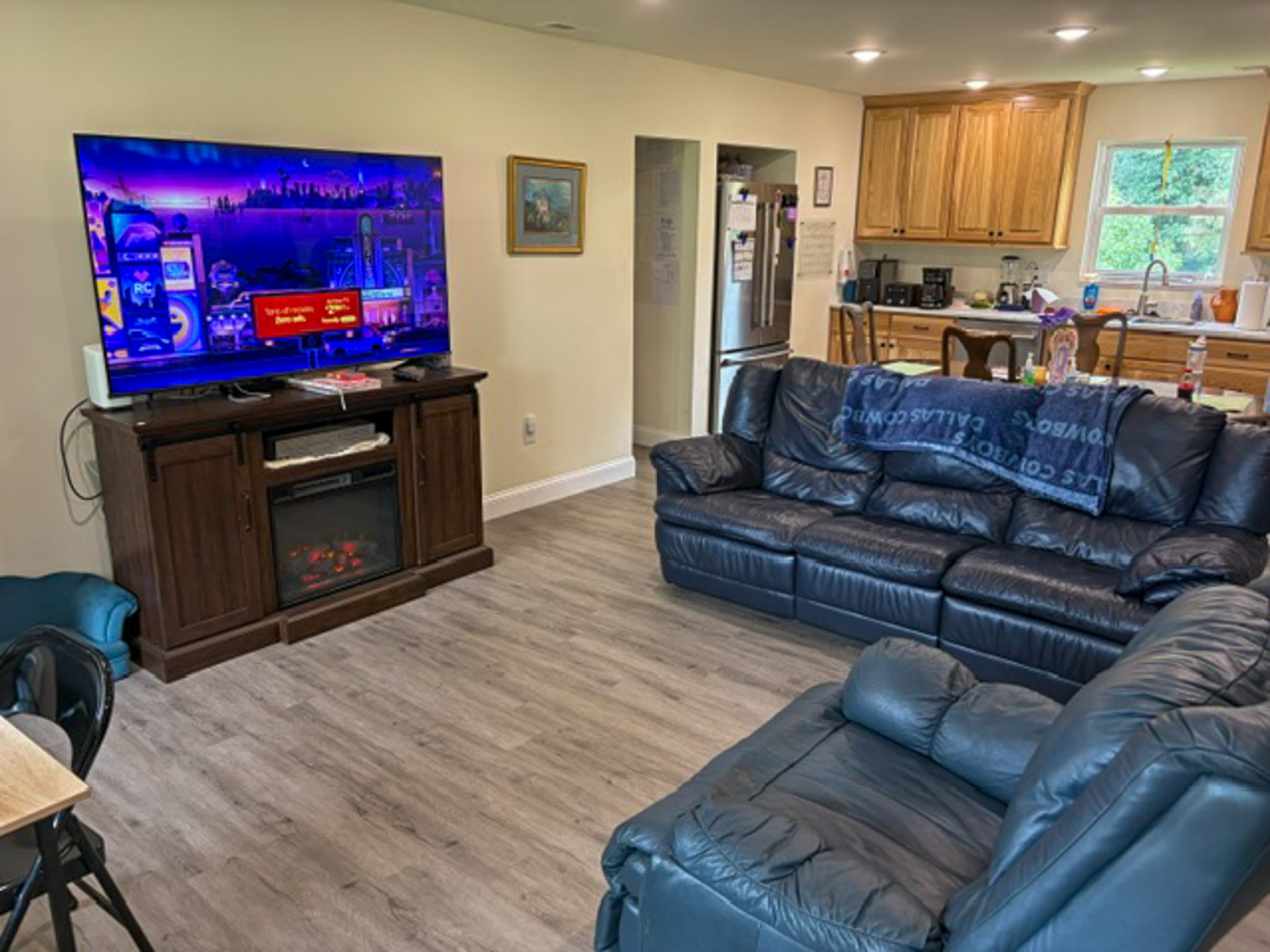 Open-concept living room featuring dark leather seating, a large television on an entertainment center with an electric fireplace, and an adjacent kitchen with natural wood cabinets, stainless steel appliances, and a dining table. Large windows provide natural light throughout the space inside the home of Group Home Provider Luke Haynes in Christiansburg, VA.