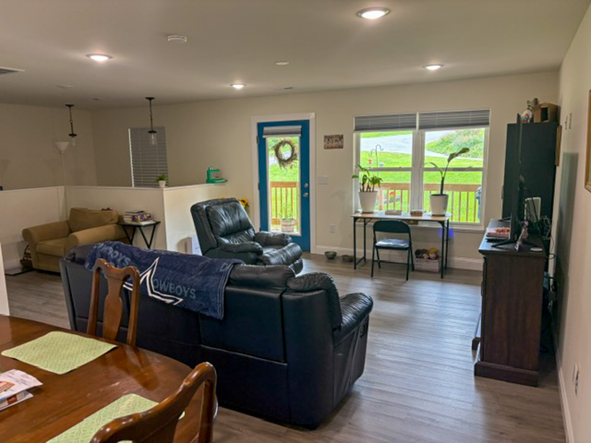 Open living area featuring black leather seating, a television console, and a small desk with potted plants positioned in front of large windows. A blue-framed glass door opens to an outdoor deck, and the space includes gray wood-look flooring, neutral walls, and recessed lighting inside the home of Group Home Provider Luke Haynes in Christiansburg, VA.