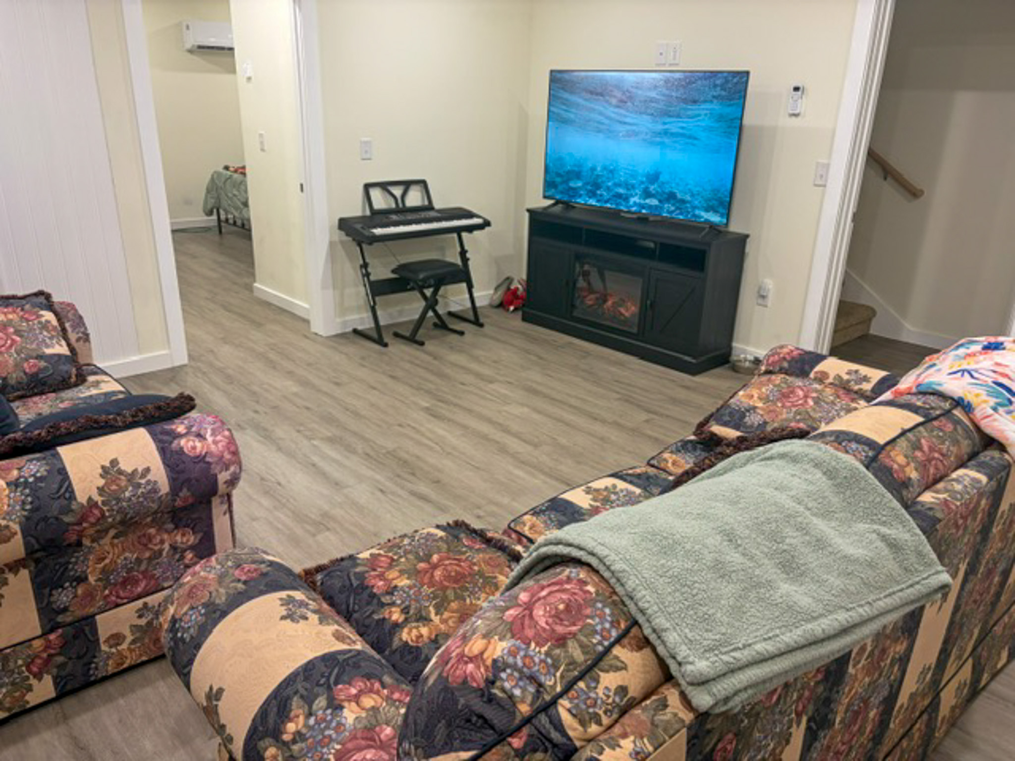 Comfortable family room with patterned sofas arranged around a television mounted on an entertainment center with an electric fireplace. The room features gray wood-look flooring, recessed lighting, a keyboard with bench, and doorways leading to adjacent rooms and a staircase inside the home of Group Home Provider Luke Haynes in Christiansburg, VA.