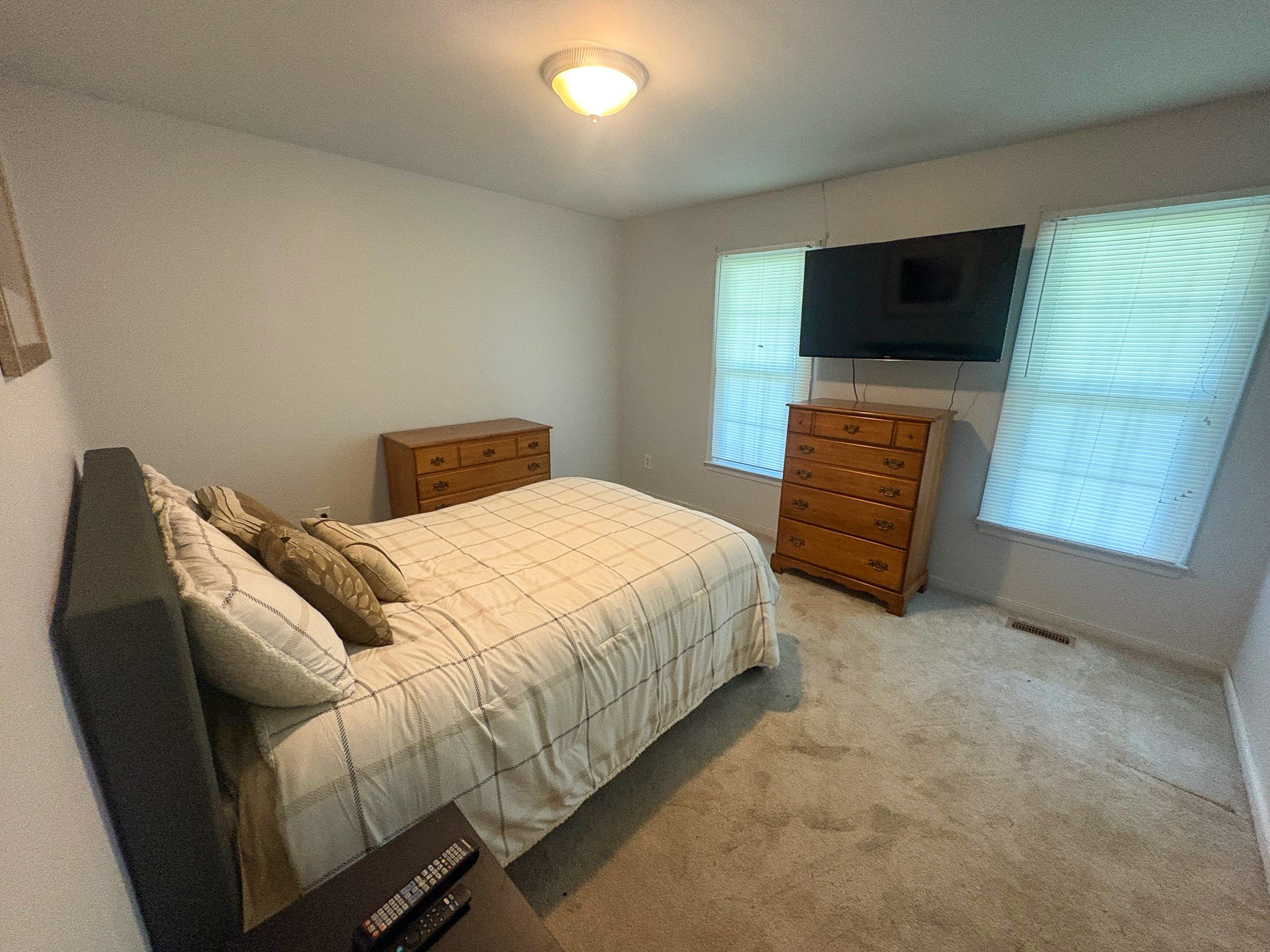 A bedroom features a neatly made bed with plaid bedding, two wooden dressers, and a wall‑mounted TV between two bright windows inside the home of Group Home Provider Lelia Mabry in Midlothian, Virginia.