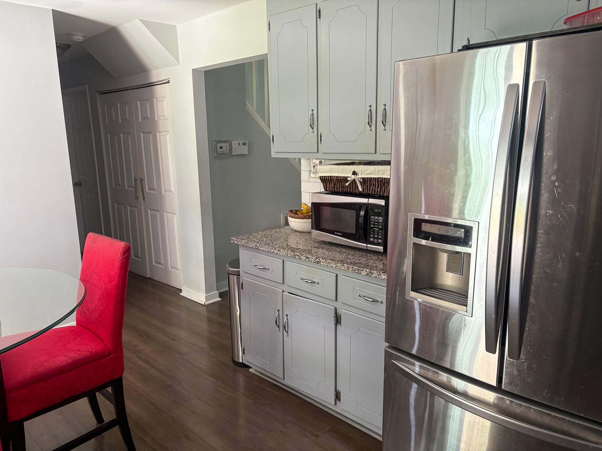 A kitchen corner features a stainless steel refrigerator beside light gray cabinets, a granite countertop, and a microwave set against the wall leading to a hallway inside the home of Group Home Provider Lelia Mabry in Midlothian, Virginia.