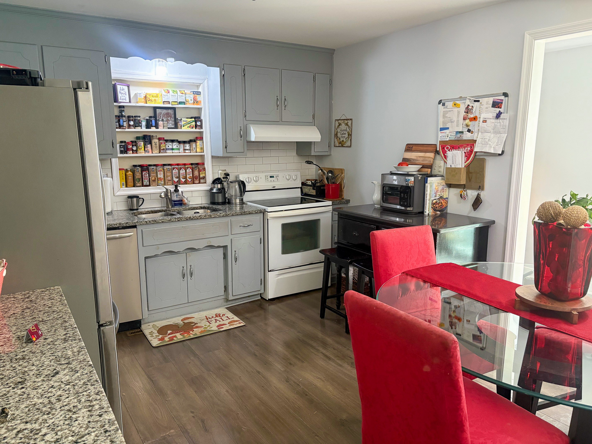 A kitchen with light gray cabinets features a white stove, open pantry shelves, and a dining area with red upholstered chairs beside a glass table inside the home of Group Home Provider Lelia Mabry in Midlothian, Virginia.