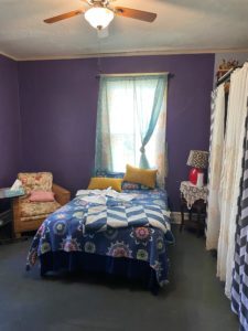 Bedroom with purple walls, a bed under a window with light curtains, an armchair on the left, and a side table with a lamp on the right inside the home of Sponsored Residential Provider Marcella Ajakaiye in Danville, Virginia.