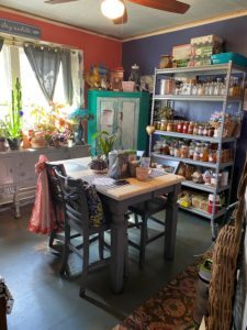 Colorful kitchen with a table and chairs in the center, shelves filled with jars and canned goods, and plants on a sunlit windowsill inside the home of Sponsored Residential Provider Marcella Ajakaiye in Danville, Virginia.