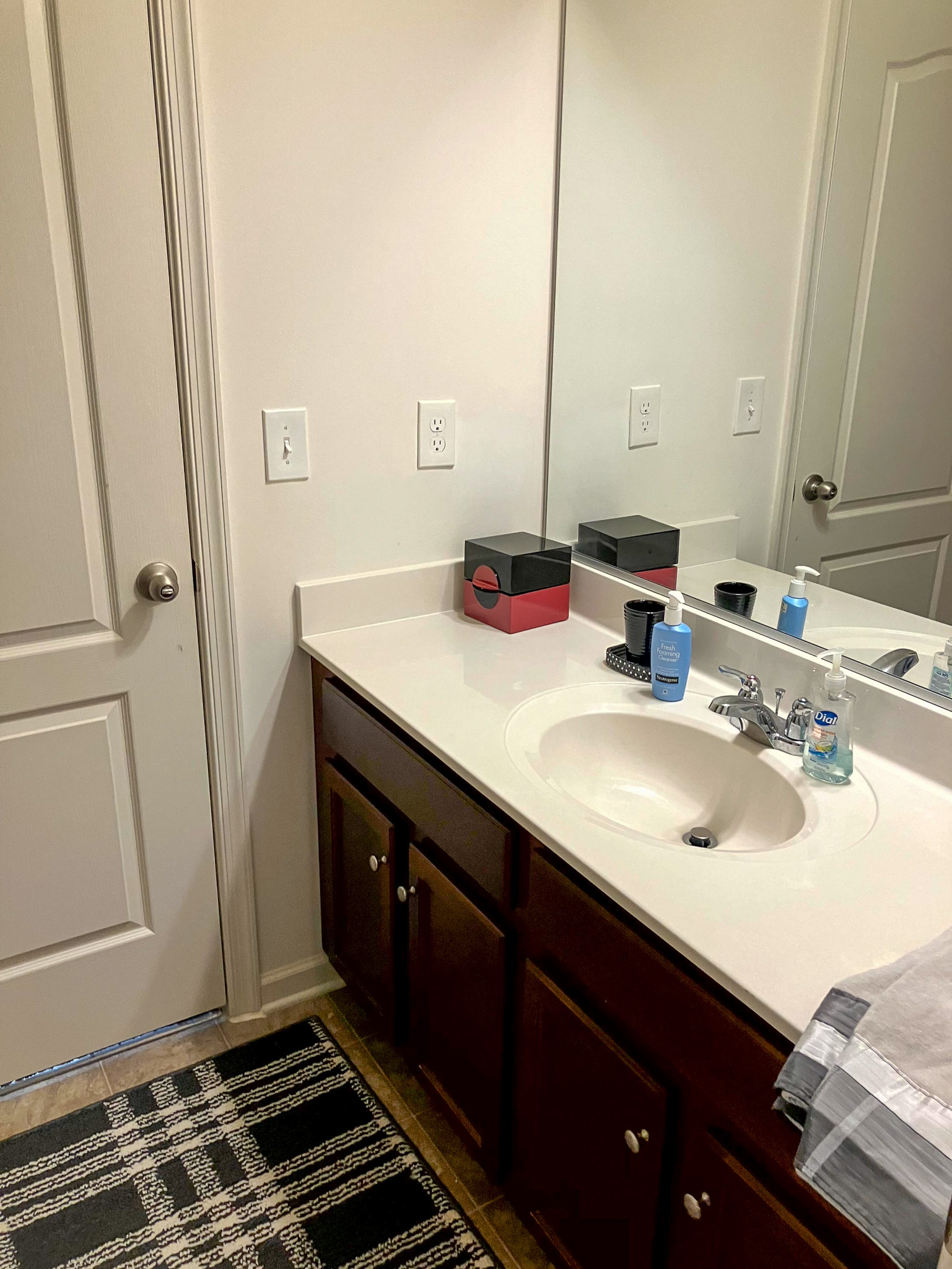 A bathroom vanity with a white countertop, a single sink, dark wood cabinets, a large mirror, and toiletries arranged on the counter inside the home of Torwon and Deljuan Mitchell in Fredericksburg, Virginia.