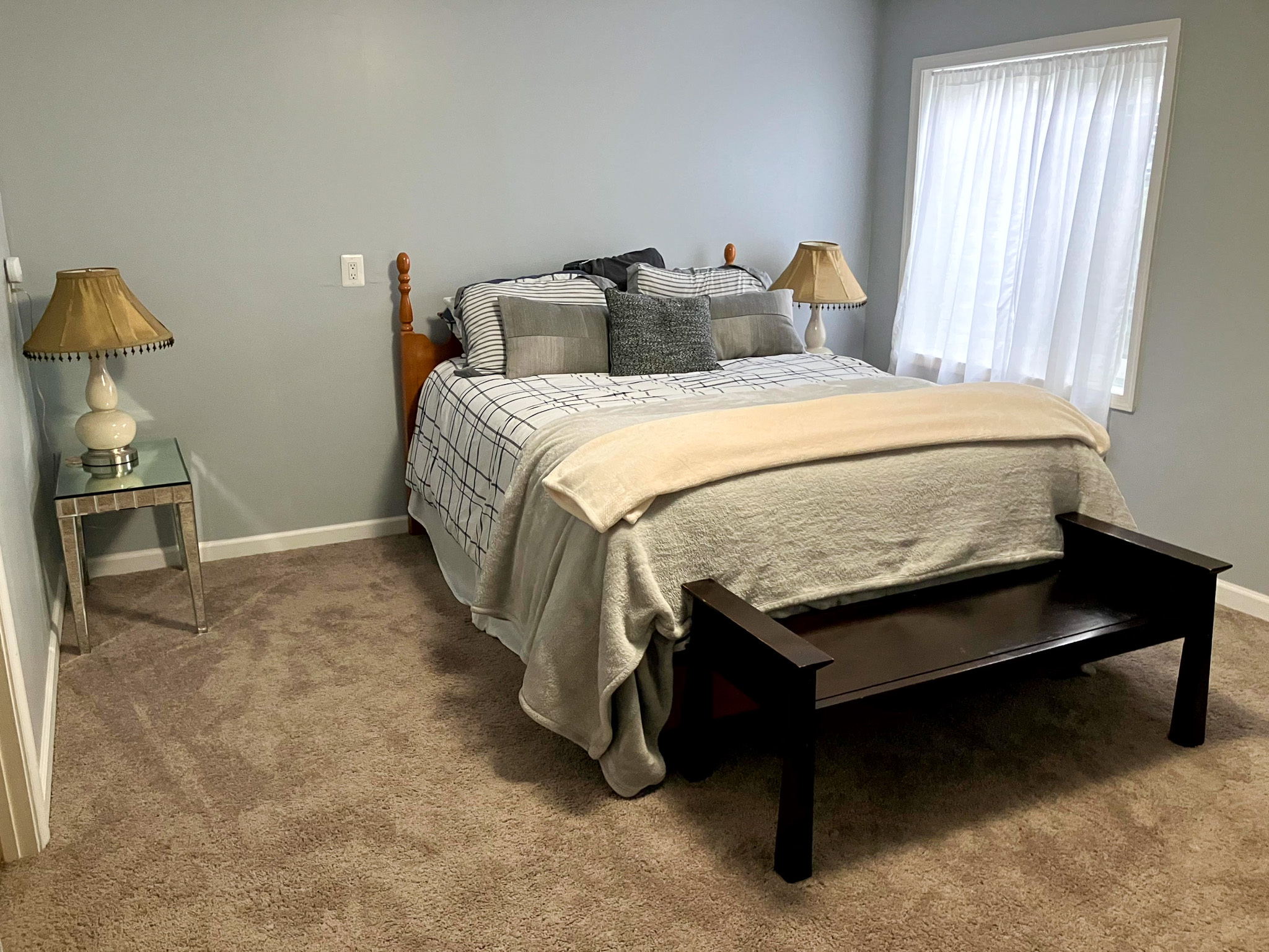 A bedroom with a neatly made bed with blue and cream bedding, matching bedside lamps, a glass-top nightstand, and a wooden bench inside the home of Torwon and Deljuan Mitchell in Fredericksburg, Virginia.