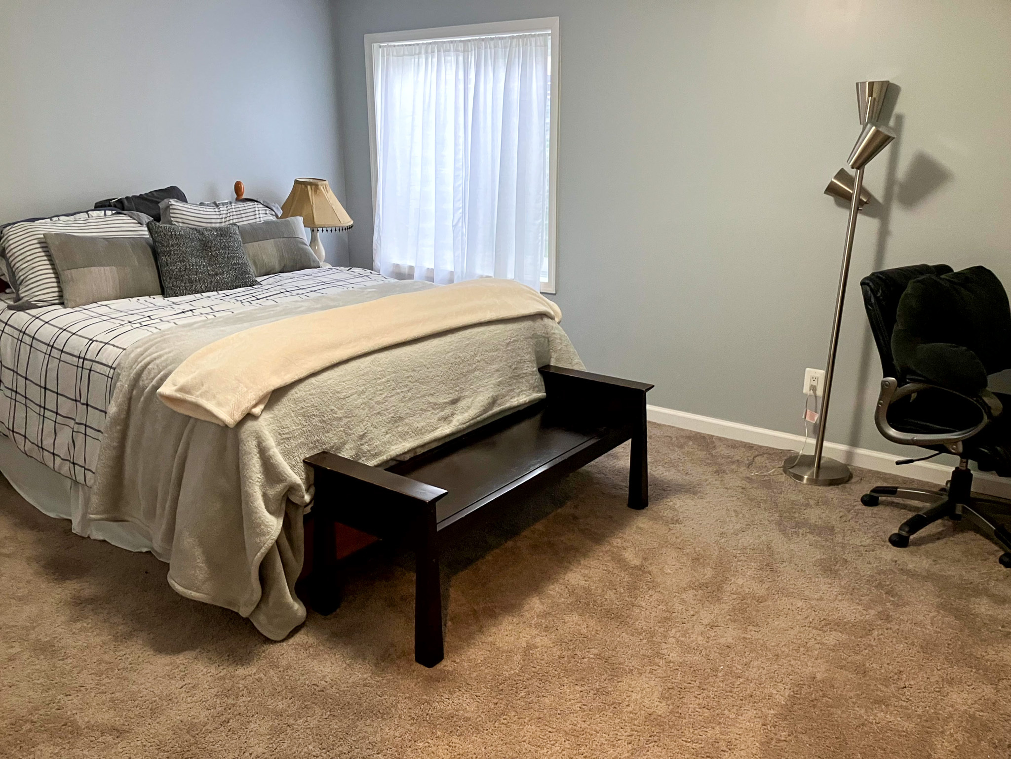 A bedroom with a neatly made bed, a wooden bench at the foot of it, a desk chair with a floor lamp, and carpeted flooring inside the home of Torwon and Deljuan Mitchell in Fredericksburg, Virginia.