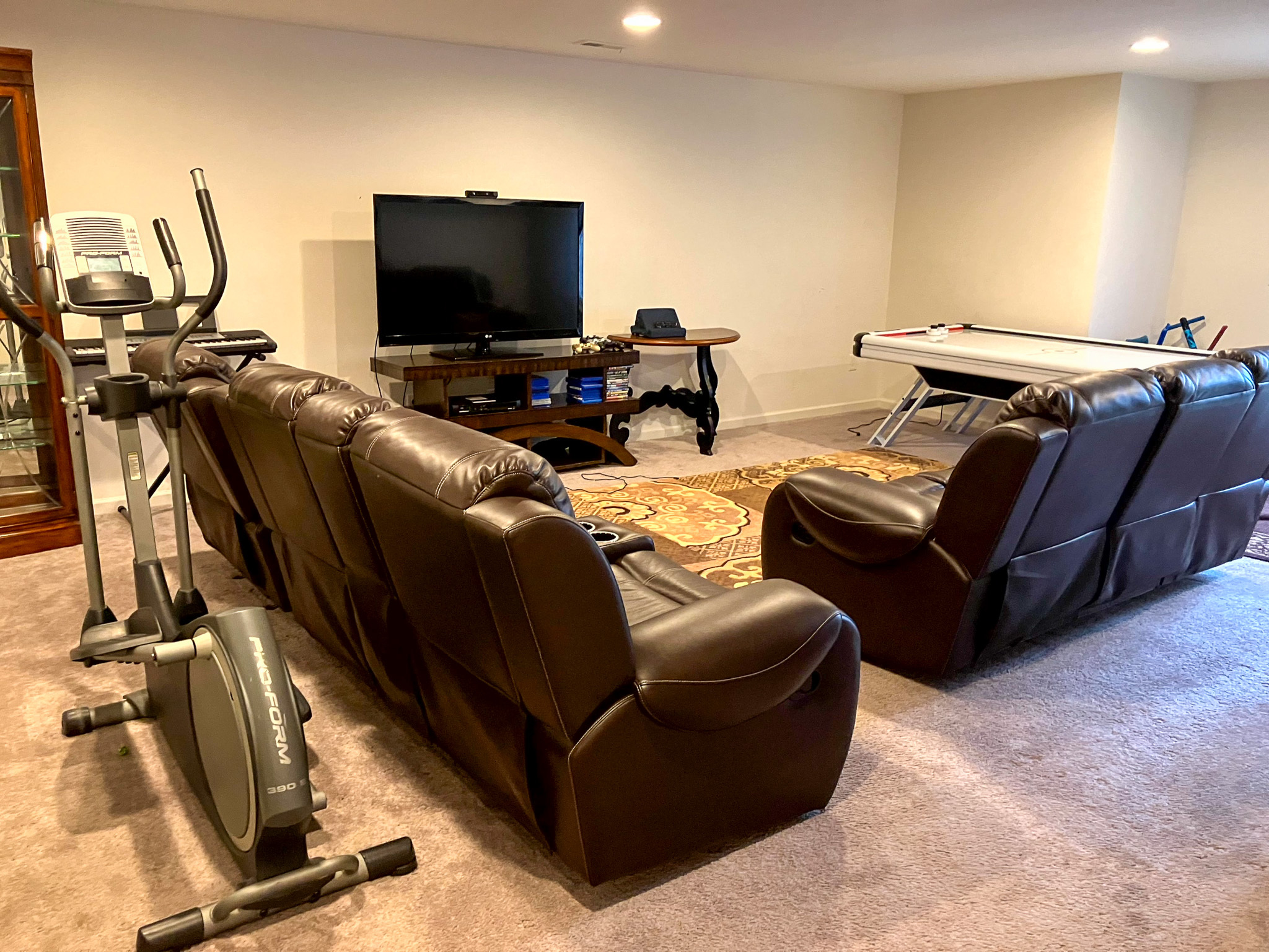A den with two brown leather sofas facing a TV, an exercise bike to the side, and an air‑hockey table in the background inside the home of Torwon and Deljuan Mitchell in Fredericksburg, Virginia.