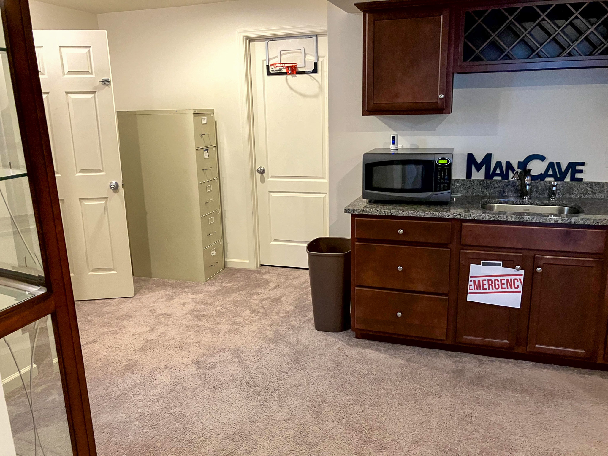 A kitchenette with dark wood cabinets, a microwave on the counter, a small sink, and a door with a mini basketball hoop with a sign that says "Man Cave" inside the home of Torwon and Deljuan Mitchell in Fredericksburg, Virginia.