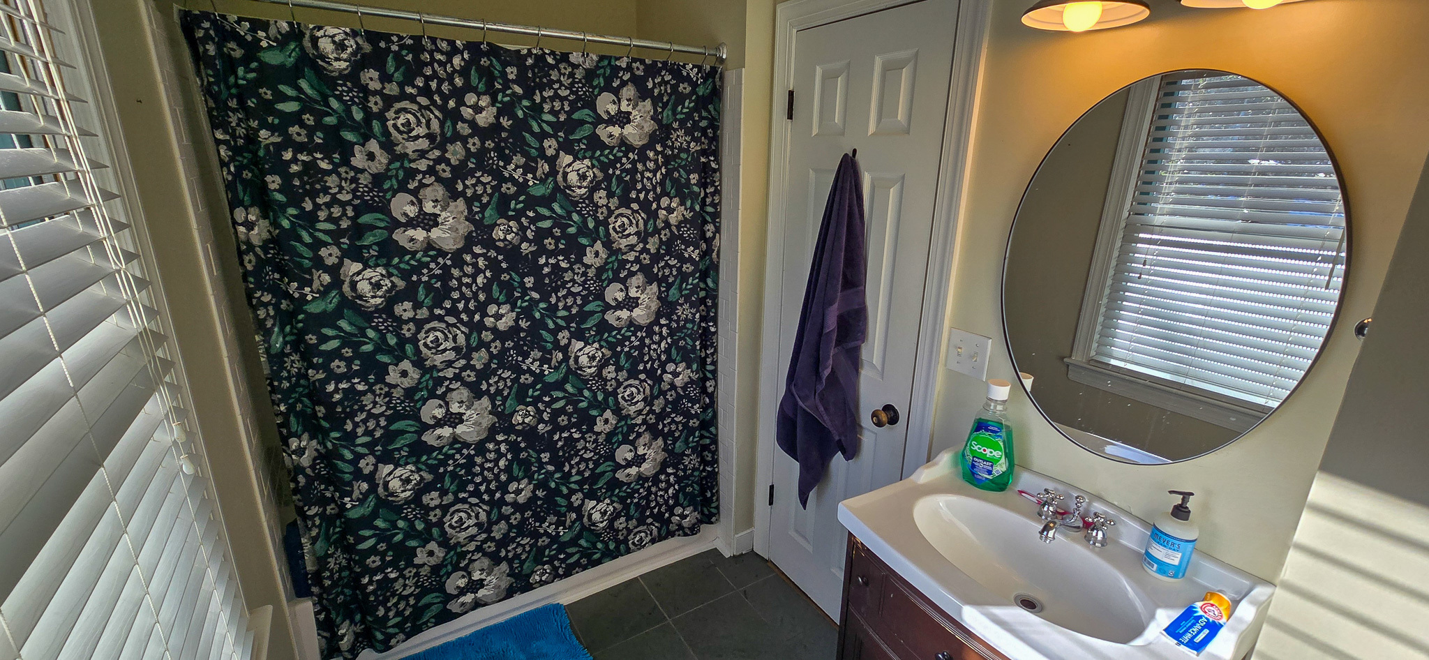 Bathroom with a round mirror above a dark vanity, window blinds, and a floral-patterned shower curtain inside the home of Group Home Provider Jared Nepal in Fredericksburg, Virginia.