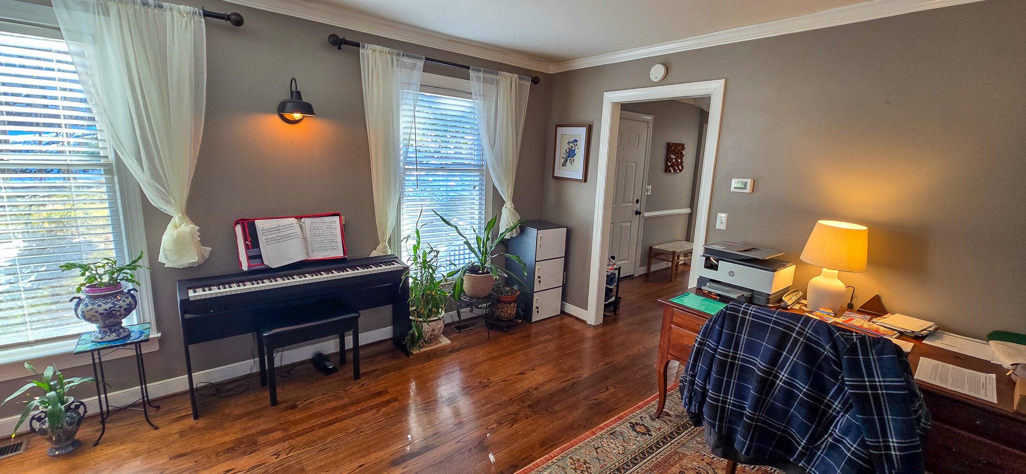 Room with a digital piano by two curtained windows, houseplants along the wall, a desk with a lamp on the right, and a doorway leading to another room inside the home of Group Home Provider Jared Nepal in Fredericksburg, Virginia.
