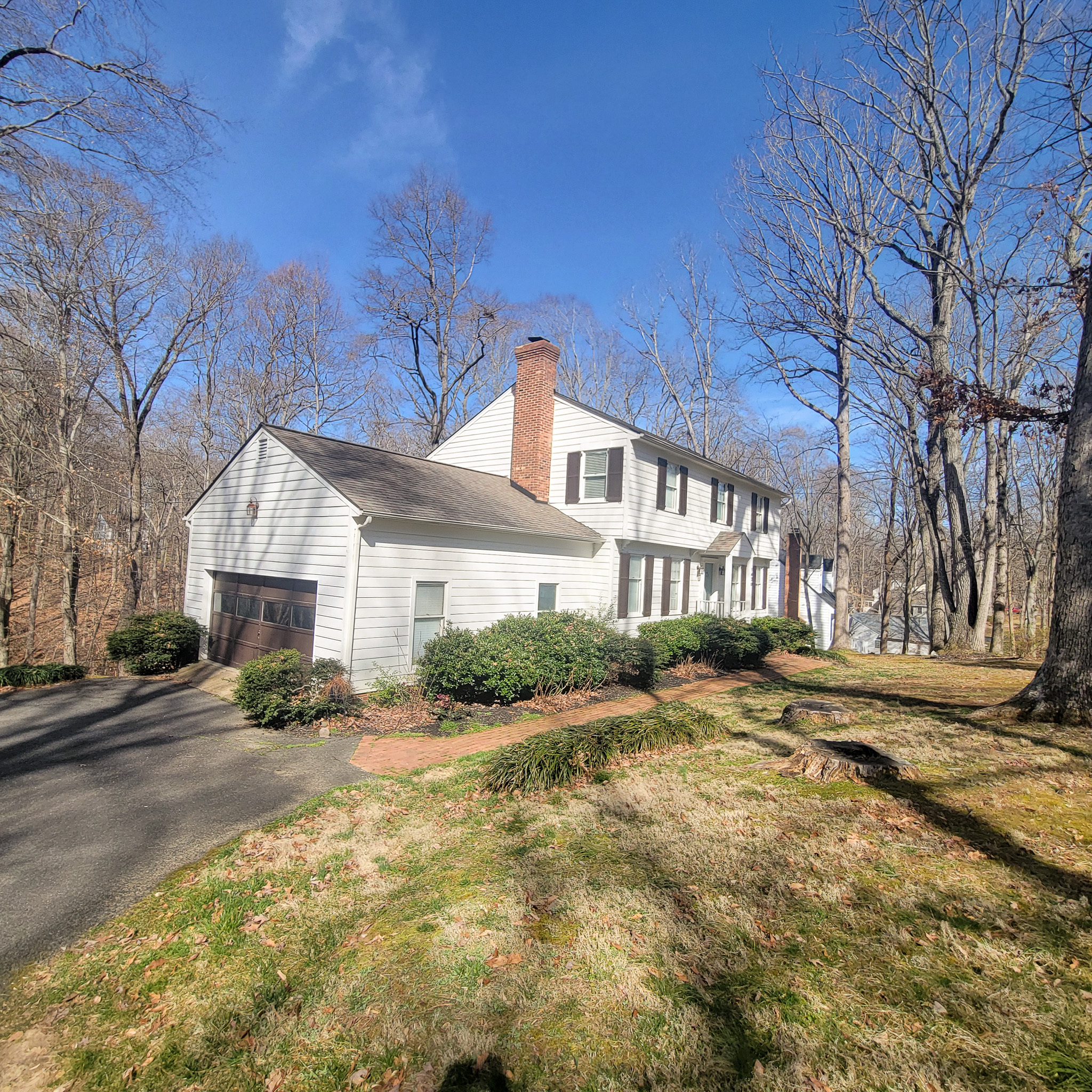 White two‑story house with black shutters, a brick chimney, and an attached garage, set on a sloped, wooded lot under a clear blue sky inside the home of Group Home Provider Jared Nepal in Fredericksburg, Virginia.