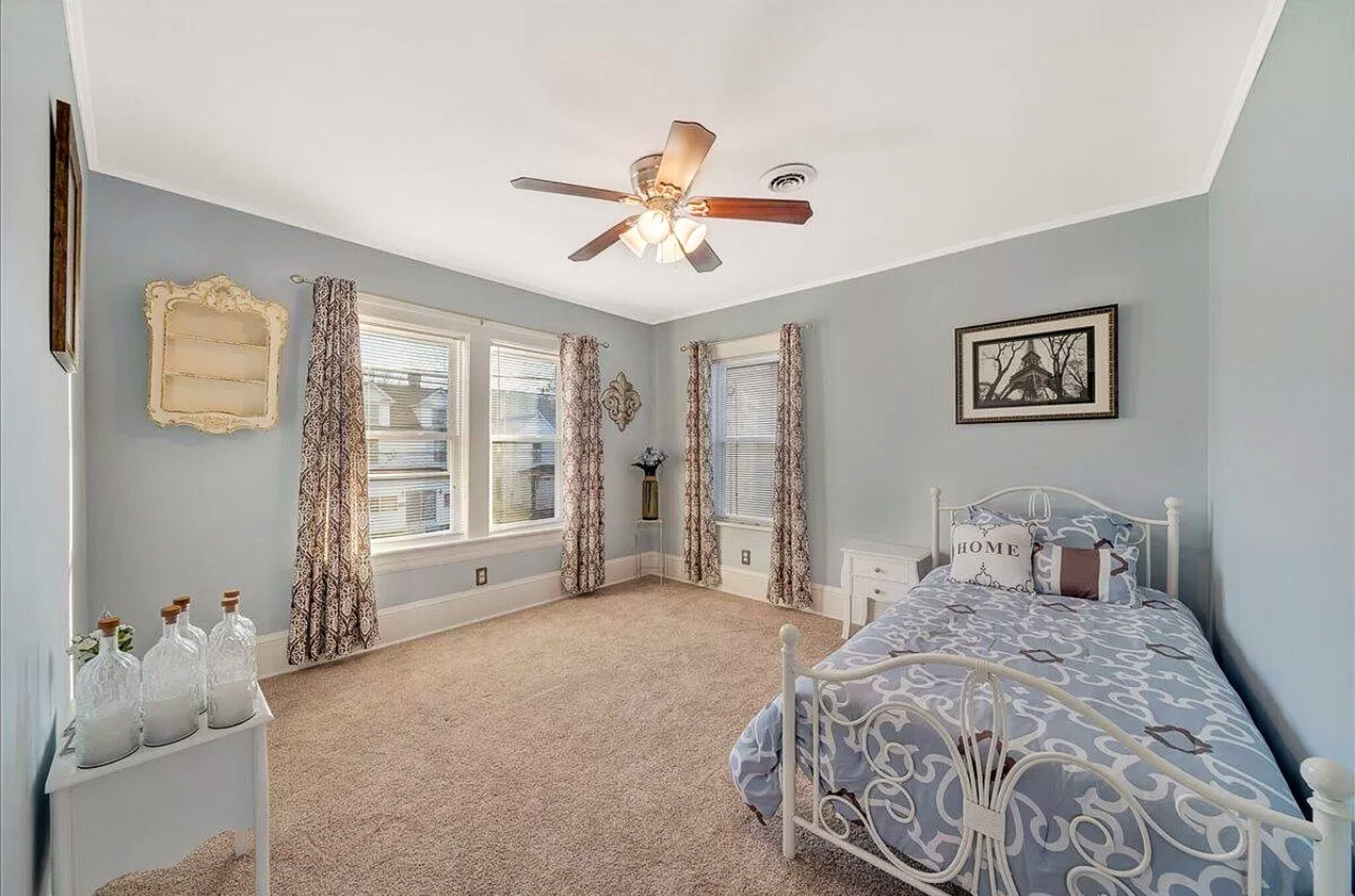 A bedroom with soft blue walls, a metal bed frame with patterned bedding, large windows with curtains, and a ceiling fan inside the home of Sponsored Residential Provider Jessica Peregrino in Roanoke, Virginia.