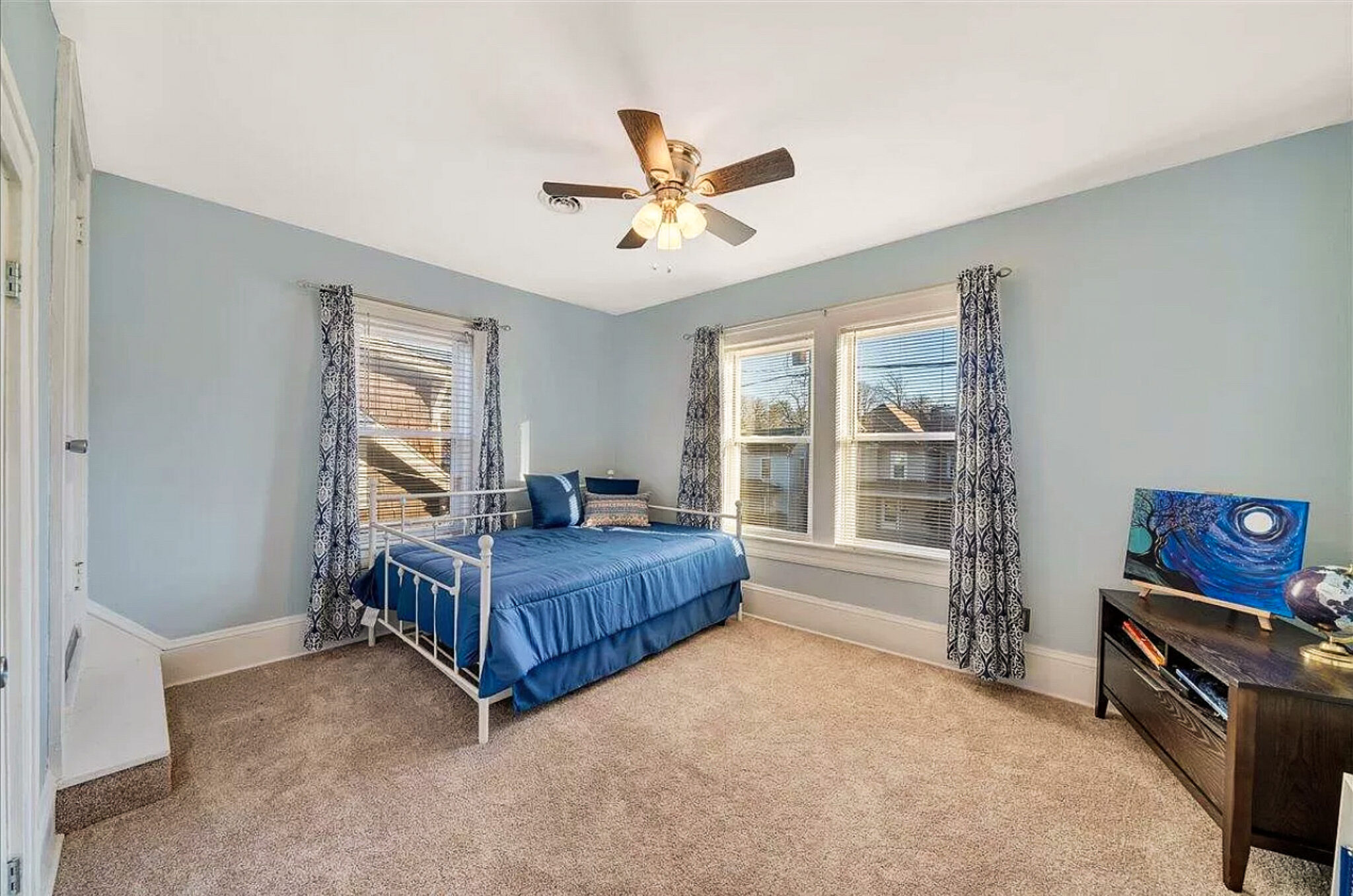 A bedroom with light blue walls, a metal bed frame with blue bedding, large windows with patterned curtains, and a ceiling fan inside the home of Sponsored Residential Provider Jessica Peregrino in Roanoke, Virginia.
