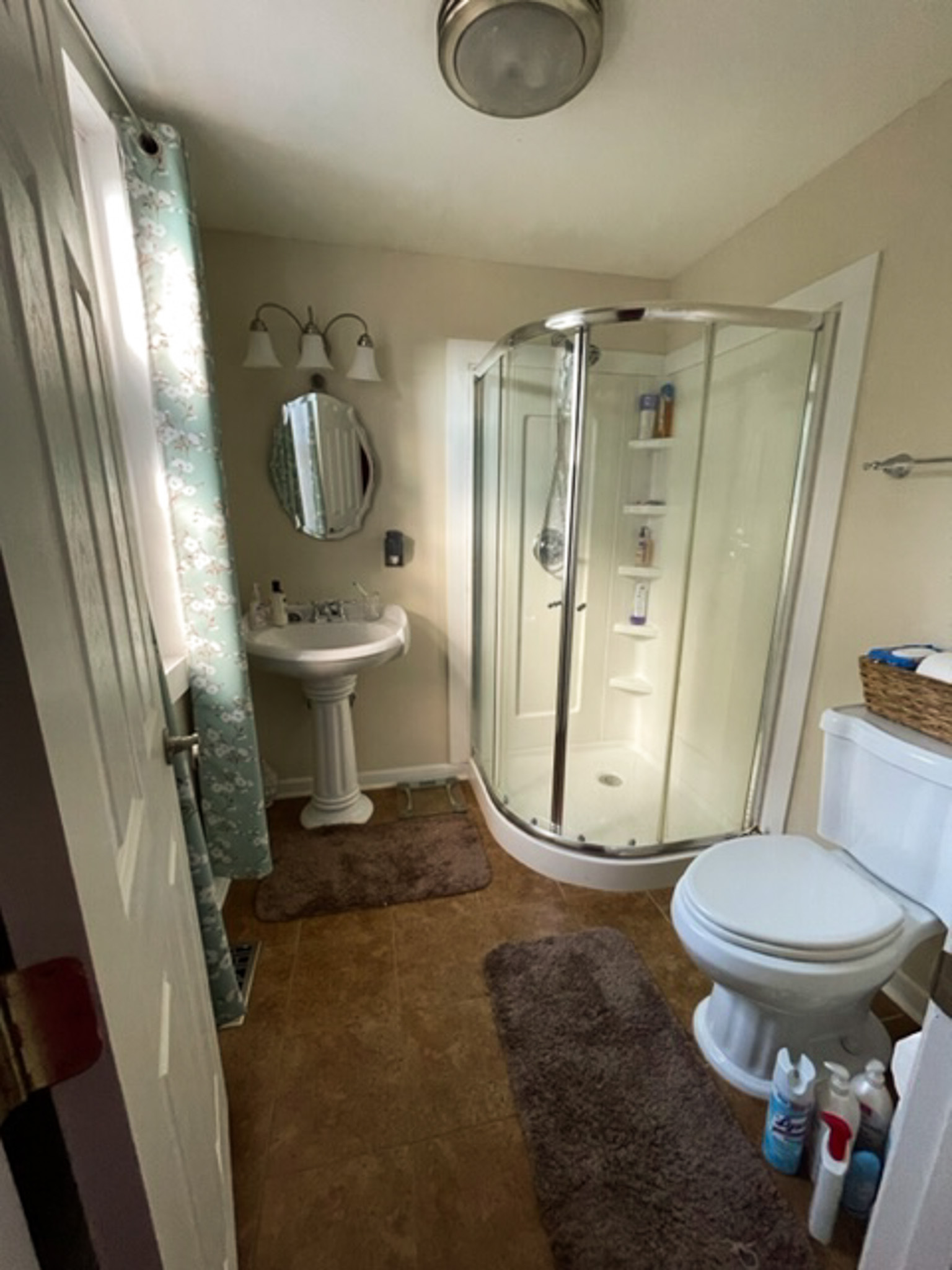 A bathroom with a corner glass shower, pedestal sink, toilet, and brown floor mats inside the home of Sponsored Residential Provider Jessica Peregrino in Roanoke, Virginia.