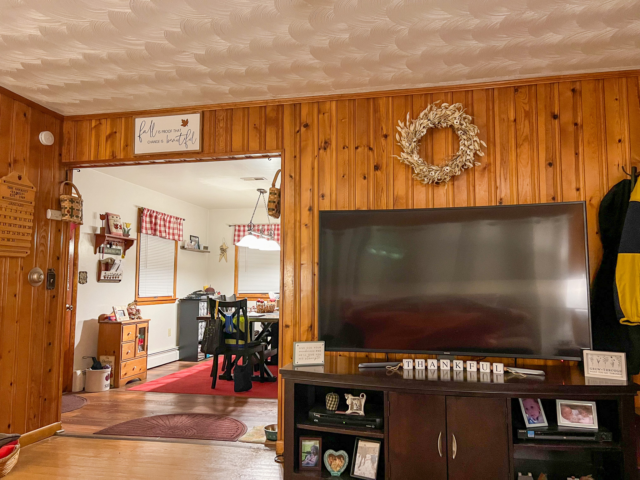 Living room with wood‑paneled walls, a large TV on a stand, and an open doorway leading to a kitchen with red accents inside the home of Sponsored Residential Provider Rebecca Sherfey in Edinburg, Virginia.
