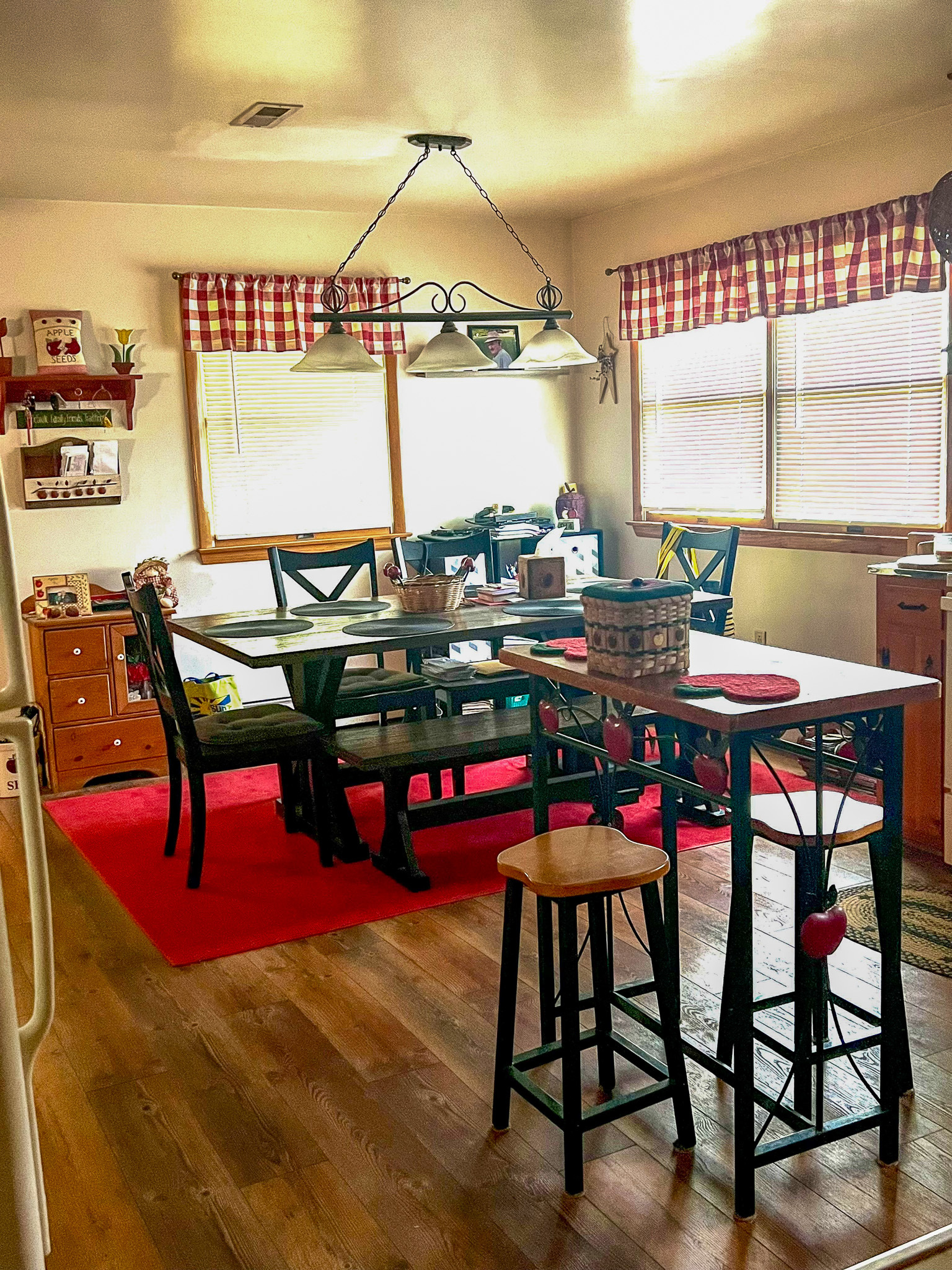Bright kitchen and dining area with wood floors, a large dining table on a red rug, checked red‑and‑white valances, and a small island with stools inside the home of Sponsored Residential Provider Rebecca Sherfey in Edinburg, Virginia.