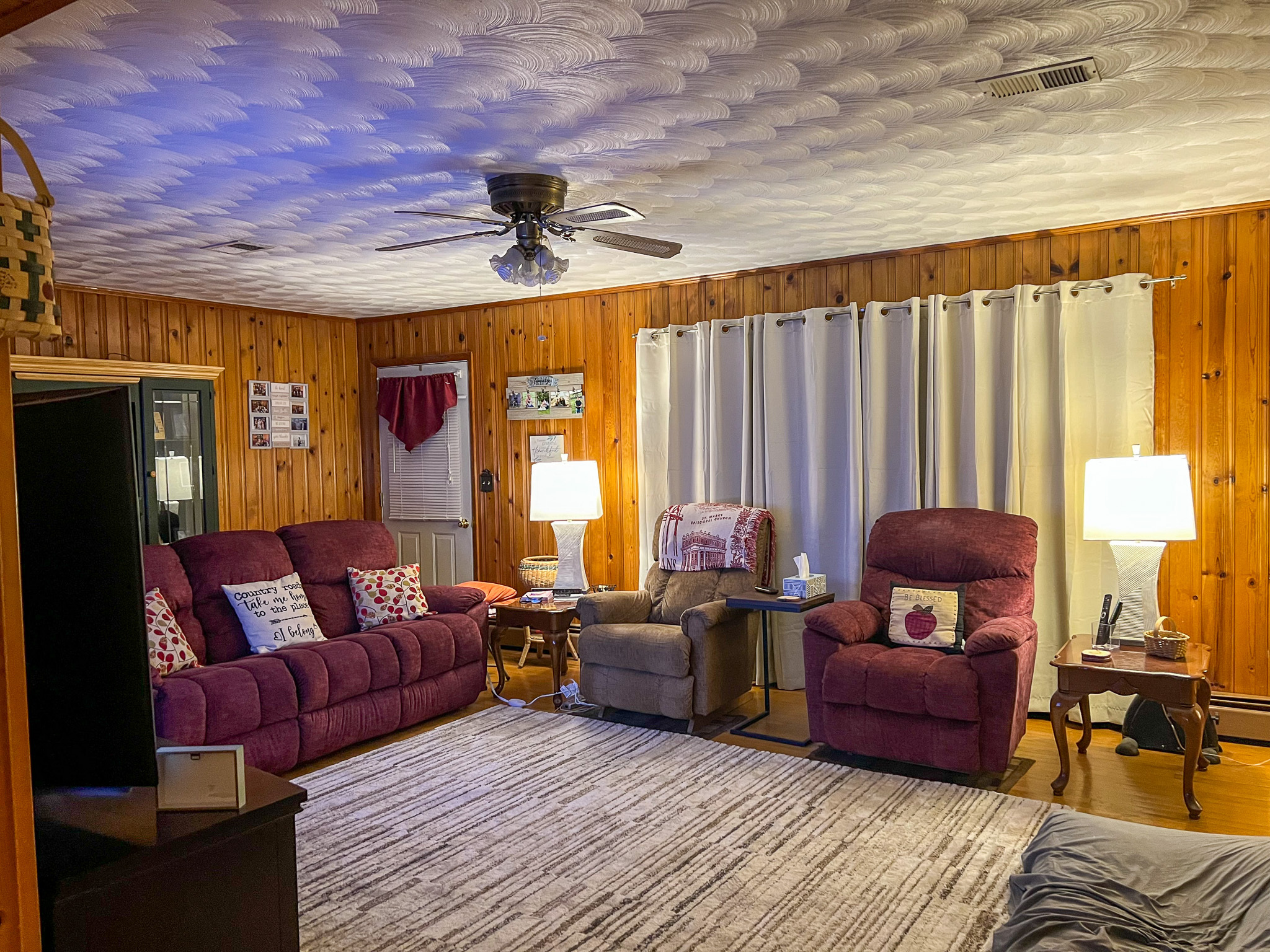 Cozy living room with wood‑paneled walls, two burgundy recliners, a matching sofa, side tables with lamps, and a large area rug beneath a ceiling fan inside the home of Sponsored Residential Provider Rebecca Sherfey in Edinburg, Virginia.