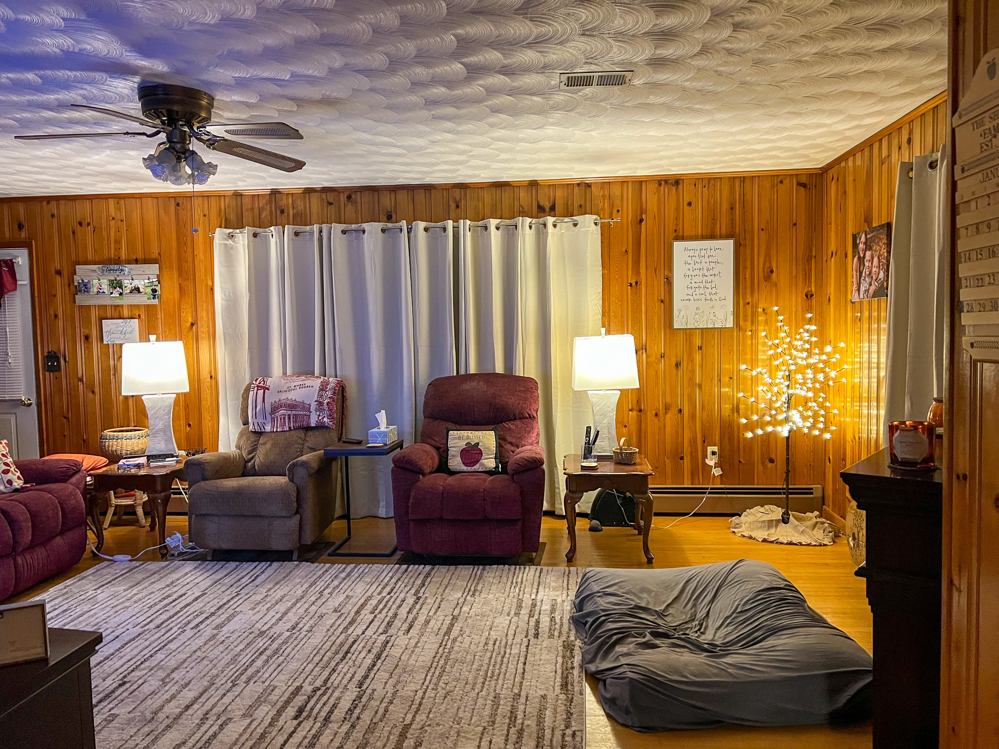 Cozy living room with wood-paneled walls, three recliners, lamps, a lit decorative tree, and a large dog bed on the floor inside the home of Sponsored Residential Provider Rebecca Sherfey in Edinburg, Virginia.