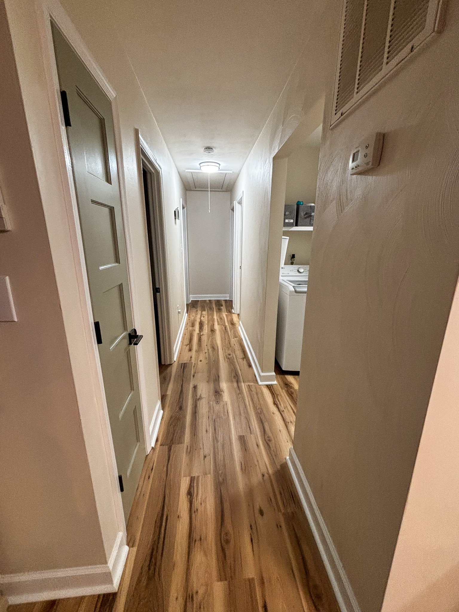 A well‑lit hallway with light wood floors, beige walls, and several doors on each side, leading to a closed door at the end inside the home of sponsored residential provider Regina Hill in Roanoke, Virginia.
