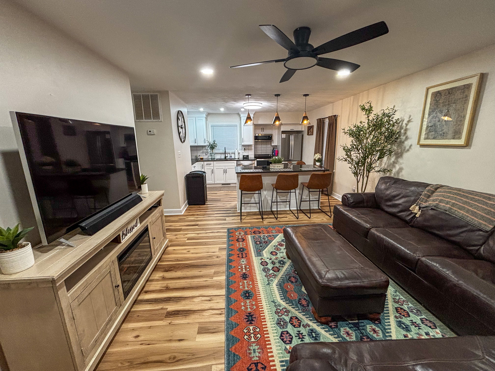 A cozy living room with a large leather sectional sofa, colorful patterned rug, and a TV on a wooden stand, opening into a bright kitchen with bar seating inside the home of sponsored residential provider Regina Hill in Roanoke, Virginia.