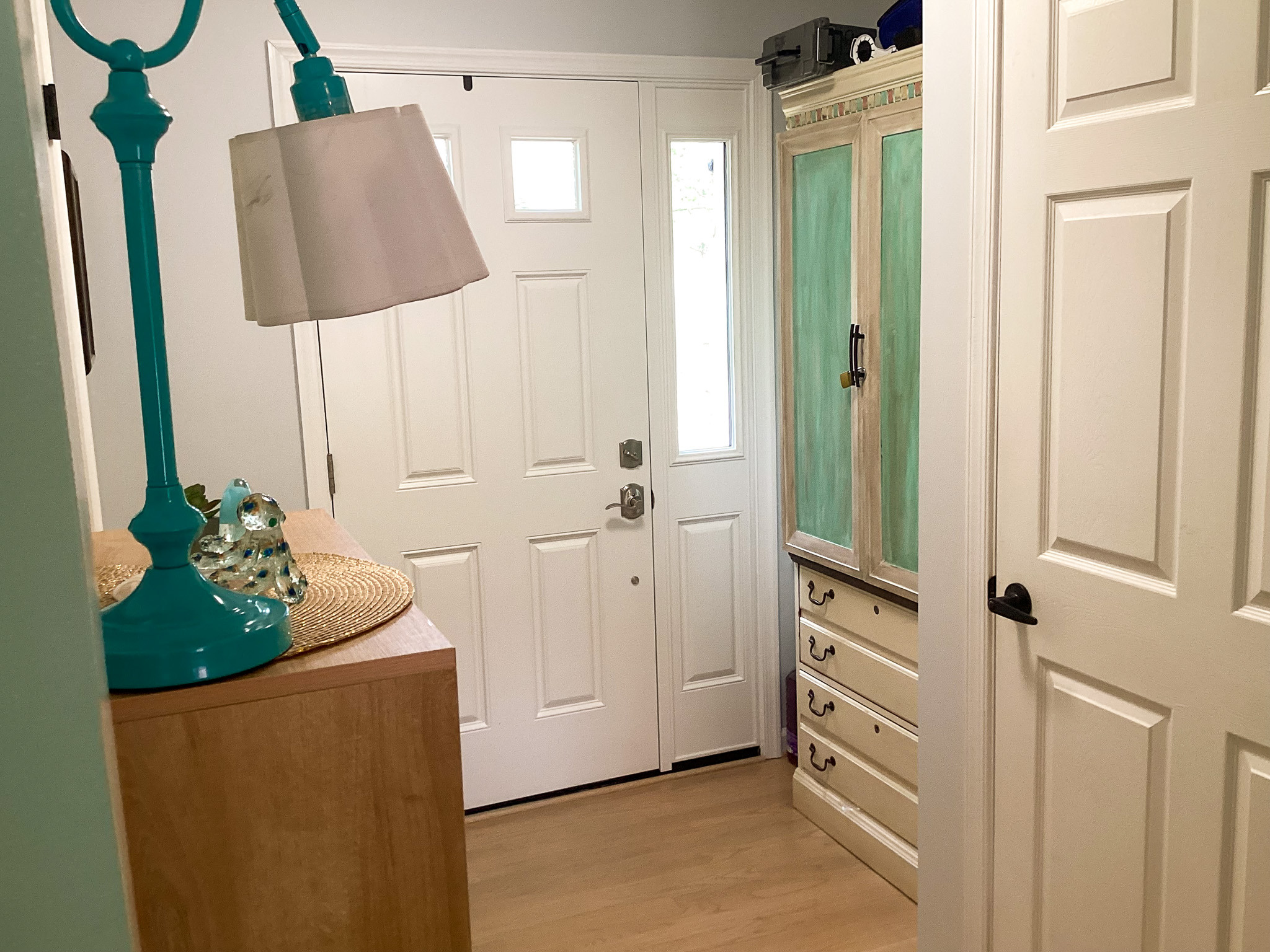 Bright, tidy entryway with a white front door, light wood cabinet, teal lamp, and accent storage, in the home of Sponsored Residential Provider Roxane Barnes.
