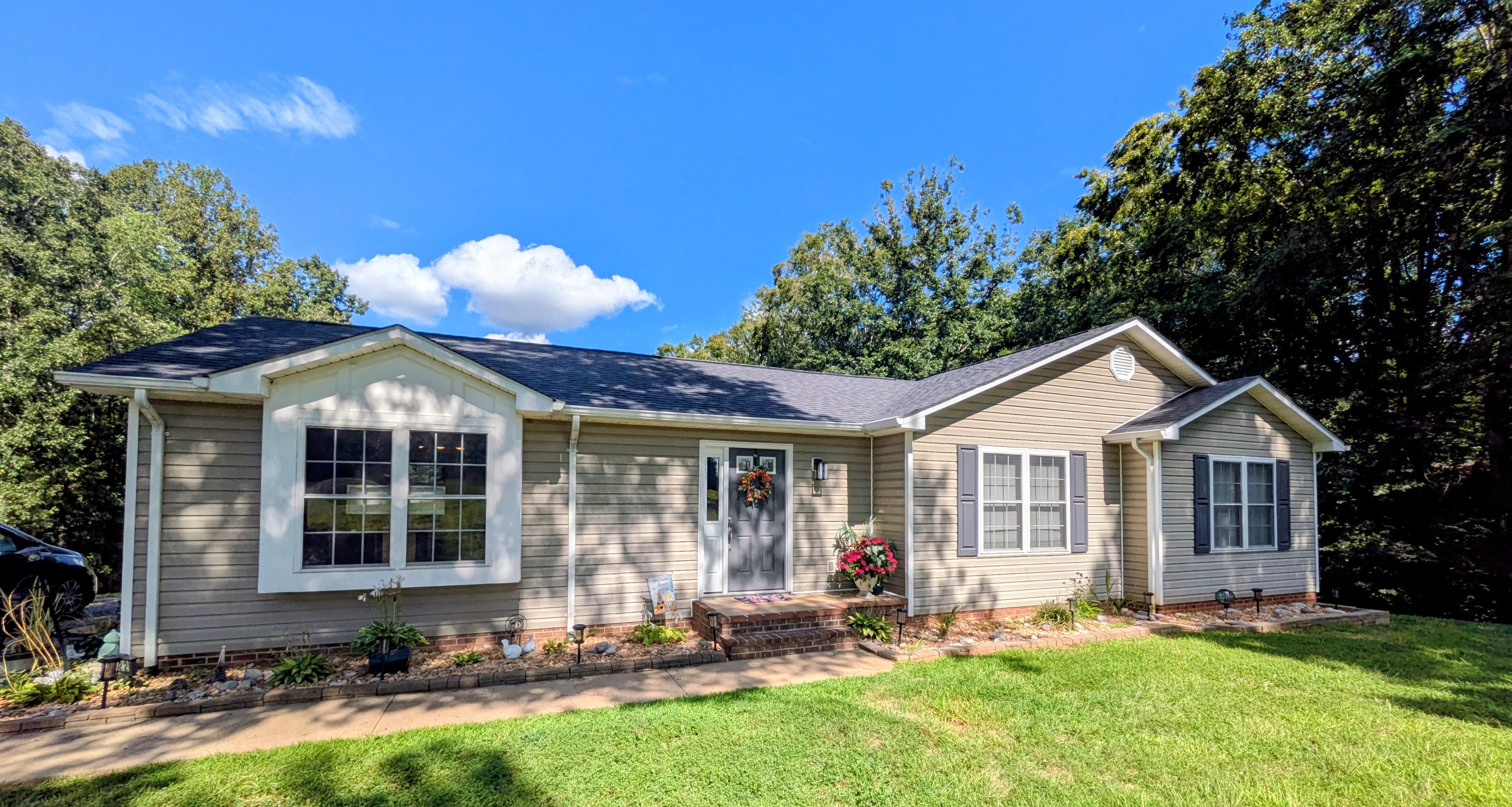 Front view of single-story beige home with dark roof, landscaped flower beds, and large lawn bordered by mature trees outside the house of Sponsored Residential Provider Roxane Barnes.