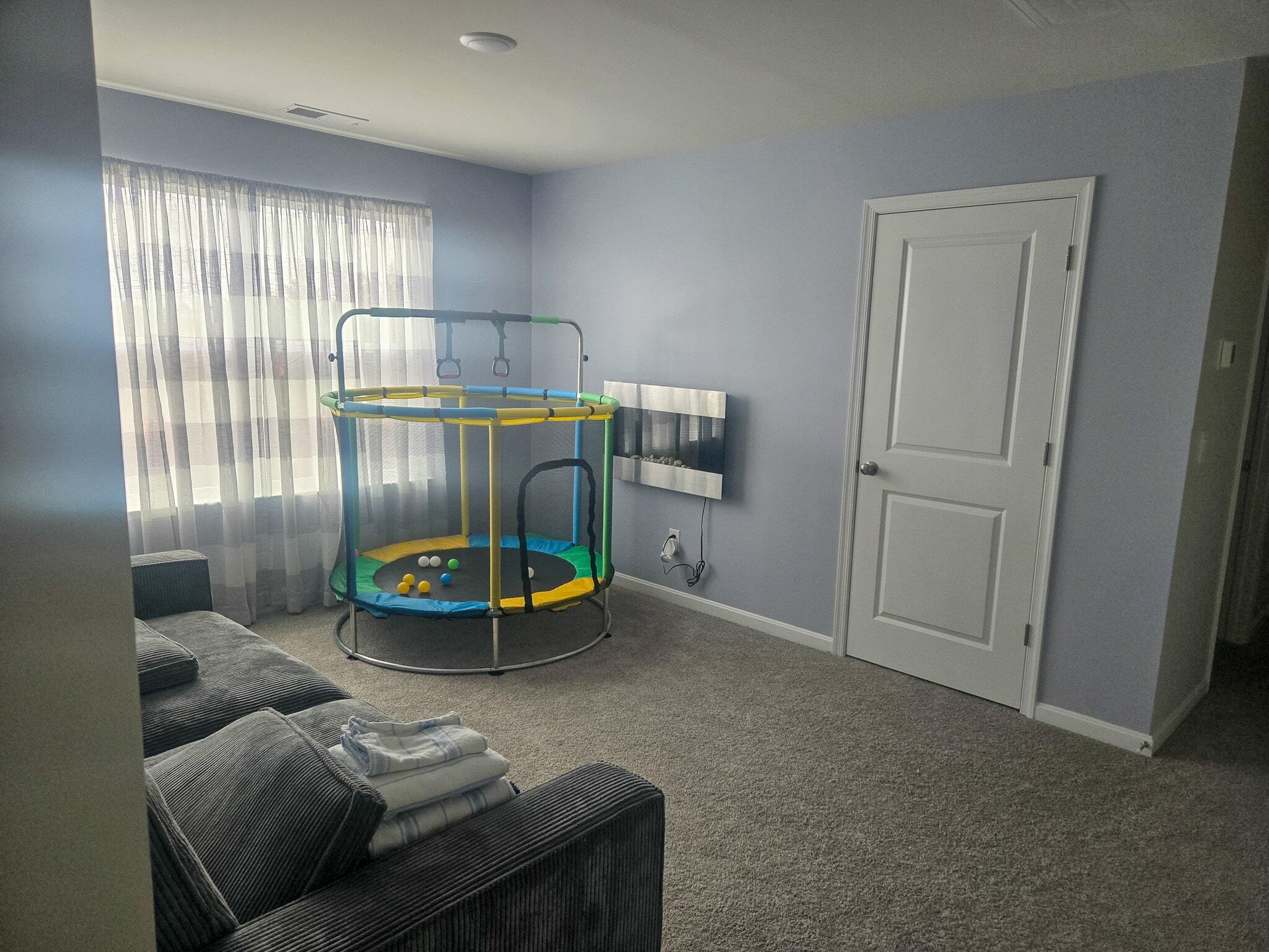 Small living area with light purple walls, a gray sofa, a mini indoor trampoline near a window with sheer curtains, and a closed white door on the opposite wall inside the home of Sponsored Residential Providers Shannon and Rebeka Knott in Staunton, Virginia.