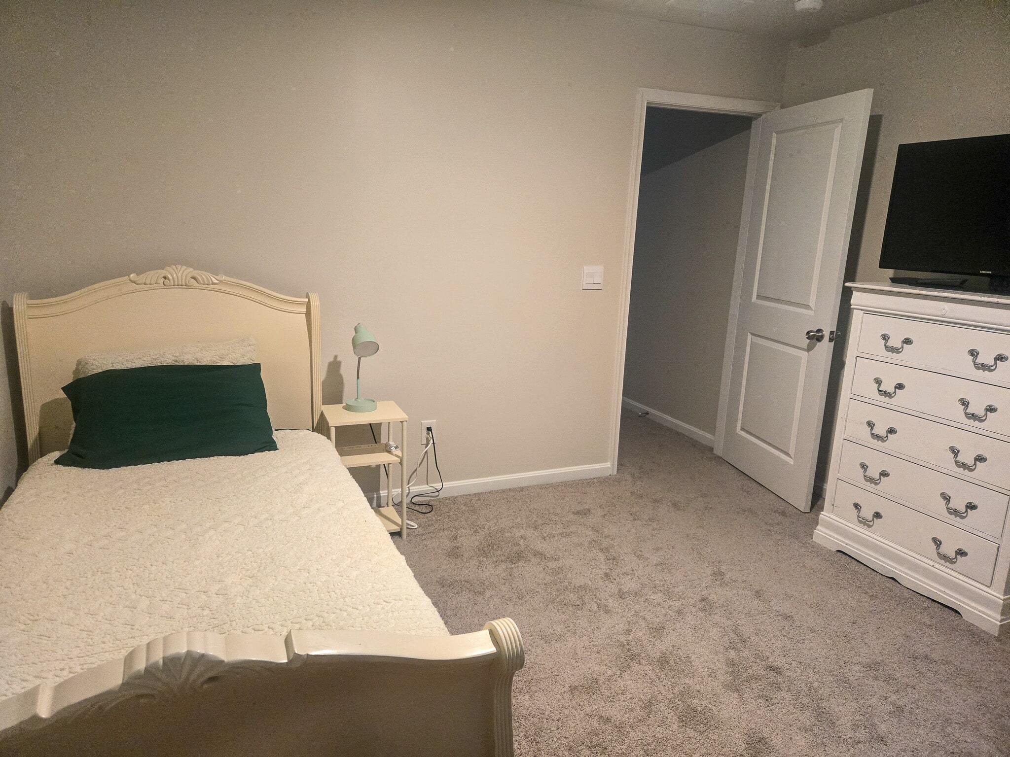 Cozy bedroom with a single bed, light-colored headboard, a green pillow, and a small bedside table with a lamp. A white dresser with a TV on top sits across the room, and an open door leads to a hallway inside the home of Sponsored Residential Providers Shannon and Rebeka Knott in Staunton, Virginia.