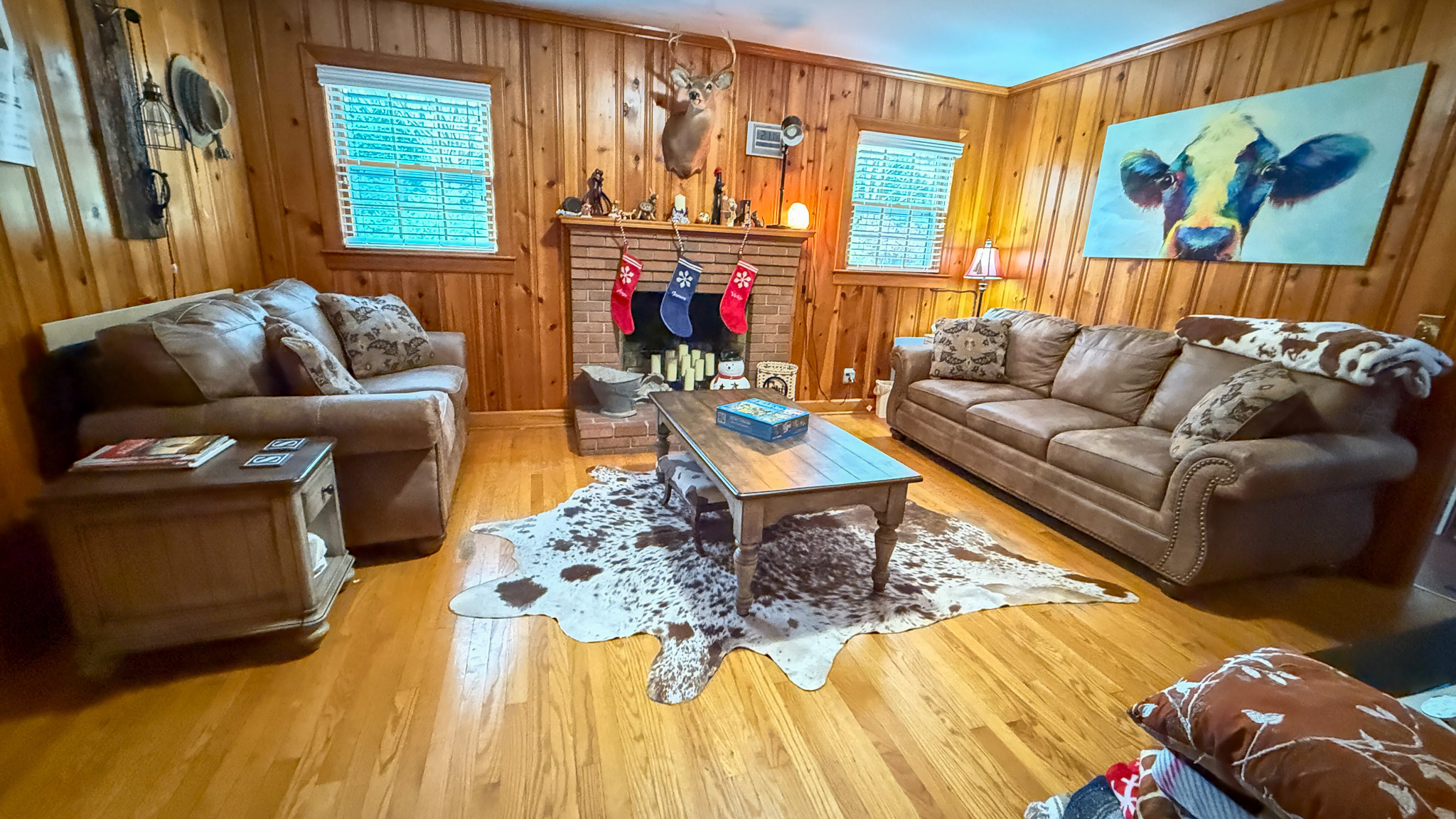A wood‑paneled living room with two sofas, a decorated fireplace, a central coffee table, and a cowhide rug on the hardwood floor inside the home of Sponsored Residential Providers Angela and James Snyder in Rocky Mount, Virginia.