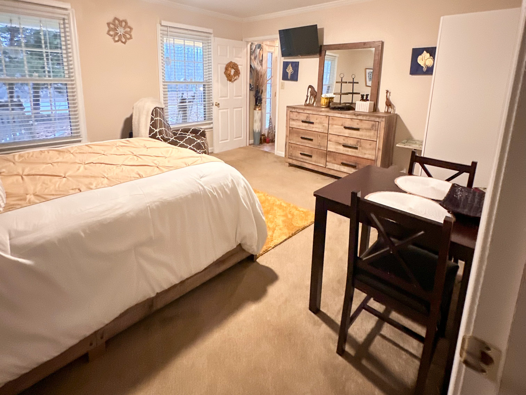 Bedroom with a wooden dresser and wall-mounted TV, small dining table set for two, and a bed with white and gold bedding inside the home of Sponsored Residential Provider Tarnisha Bradshaw in Fredericksburg, Virginia.