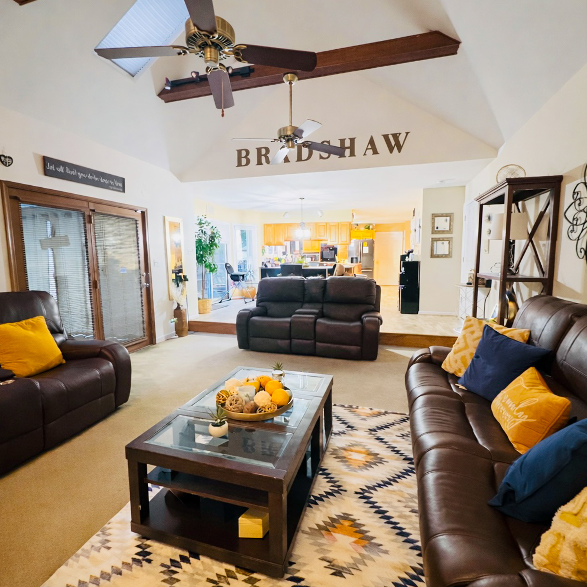 A living room with brown leather sofas, a patterned rug, a glass‑top coffee table, and exposed ceiling beams, opening into a brightly lit kitchen area inside the home of Sponsored Residential Provider Tarnisha Bradshaw in Fredericksburg, Virginia.