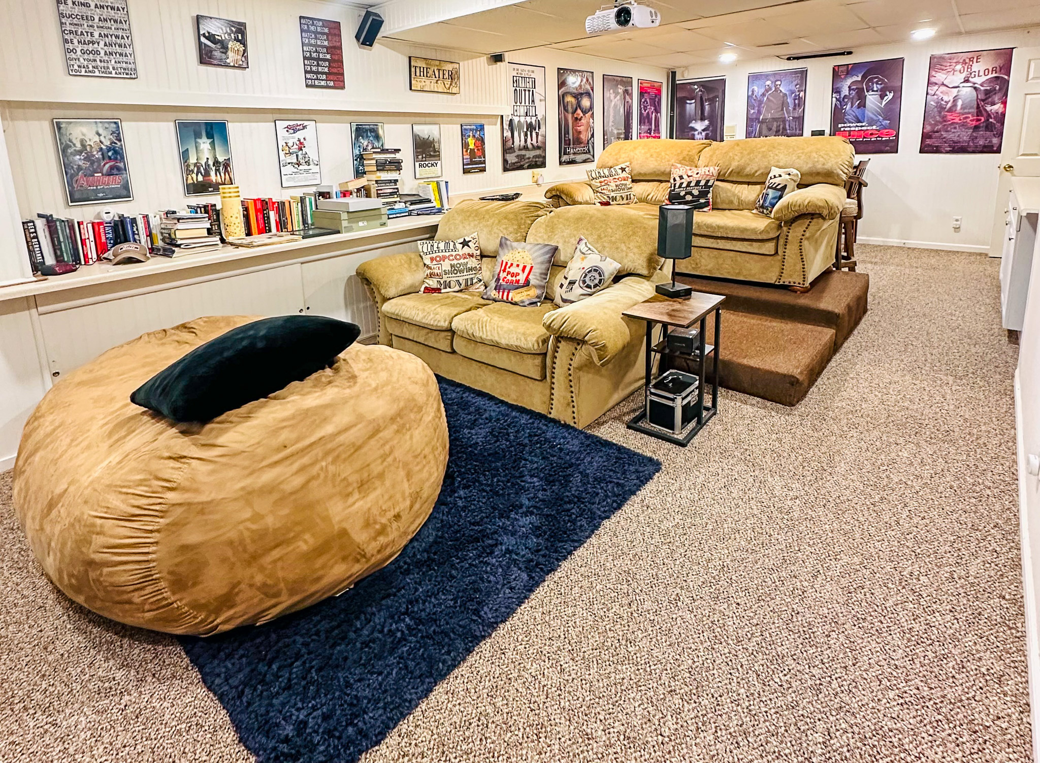 A home theater room with tiered tan recliners, a large tan beanbag chair on a blue rug, movie posters on the walls, and books and decor along built‑in shelves inside the home of Sponsored Residential Provider Tarnisha Bradshaw in Fredericksburg, Virginia
.