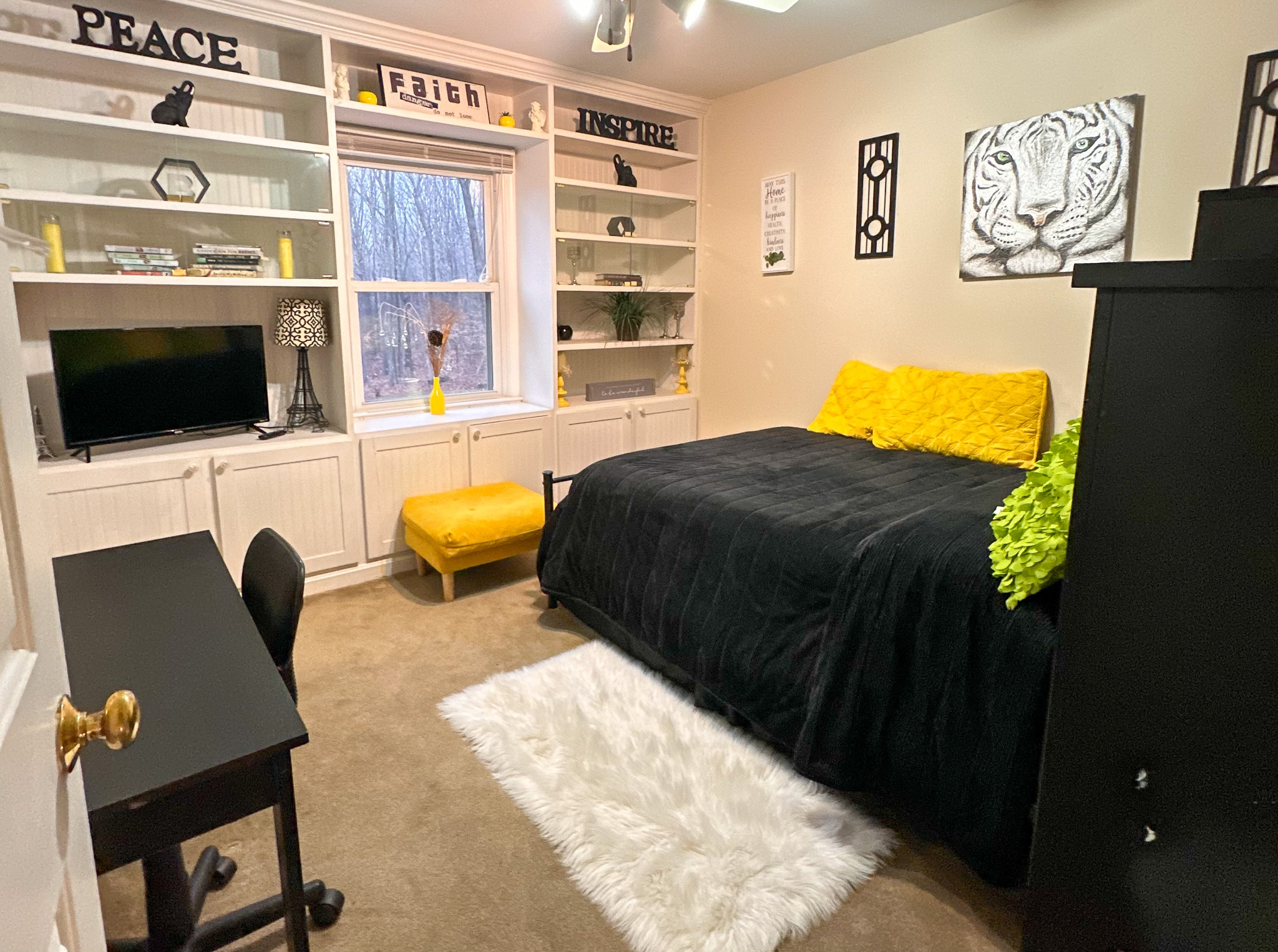 A bedroom with built‑in shelves, a bed with black bedding and yellow accents, a white rug, a desk, and a window centered between shelving units inside the home of Sponsored Residential Provider Tarnisha Bradshaw  in Fredericksburg, Virginia.