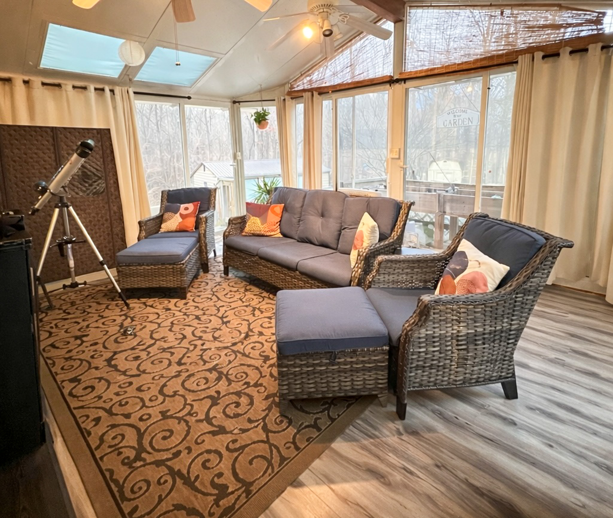 A bright sunroom with wicker furniture, blue cushions, a patterned rug, large windows, and a telescope near the wall inside the home of Sponsored Residential Provider Tarnisha Bradshaw  in Fredericksburg, Virginia.