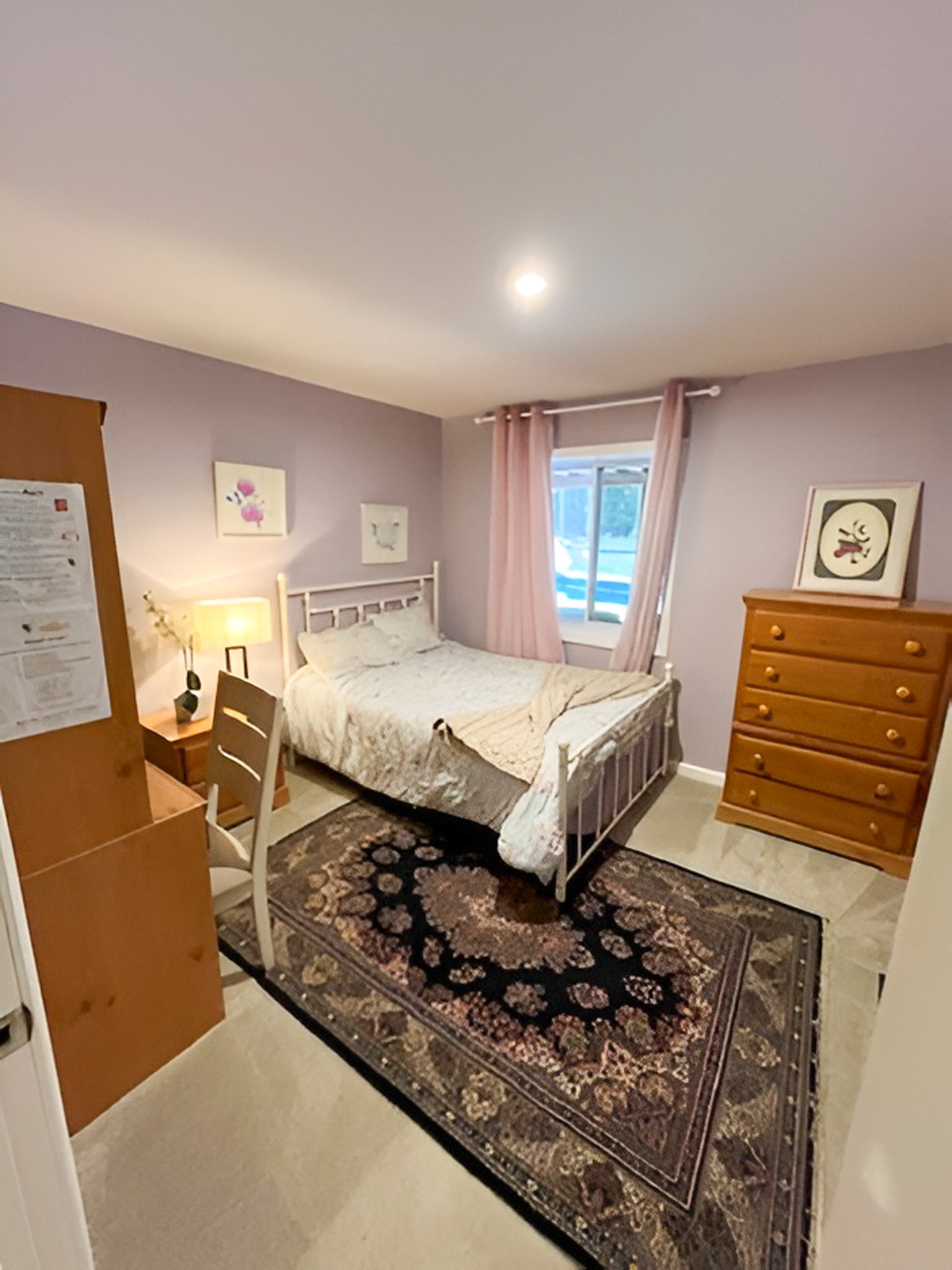 Cozy bedroom with lavender walls, a bed centered on a patterned rug, wooden dressers, and pink curtains by the window inside the home of Group Home Providers Alex and Dillon Tittle in Fredericksburg, Virginia.