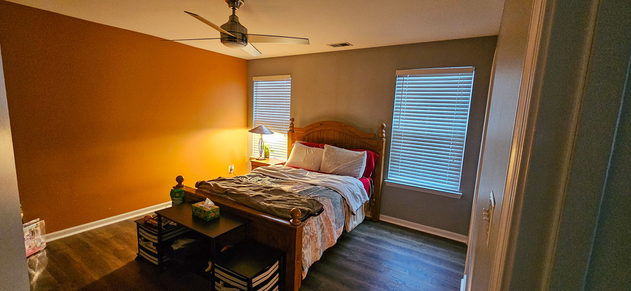 A bedroom with an orange accent wall, a wooden bed with patterned bedding, two windows, and a small bench at the foot of the bed inside the home of Sponsored Residential Provider Vanessa Moore in Fredericksburg, Virginia.